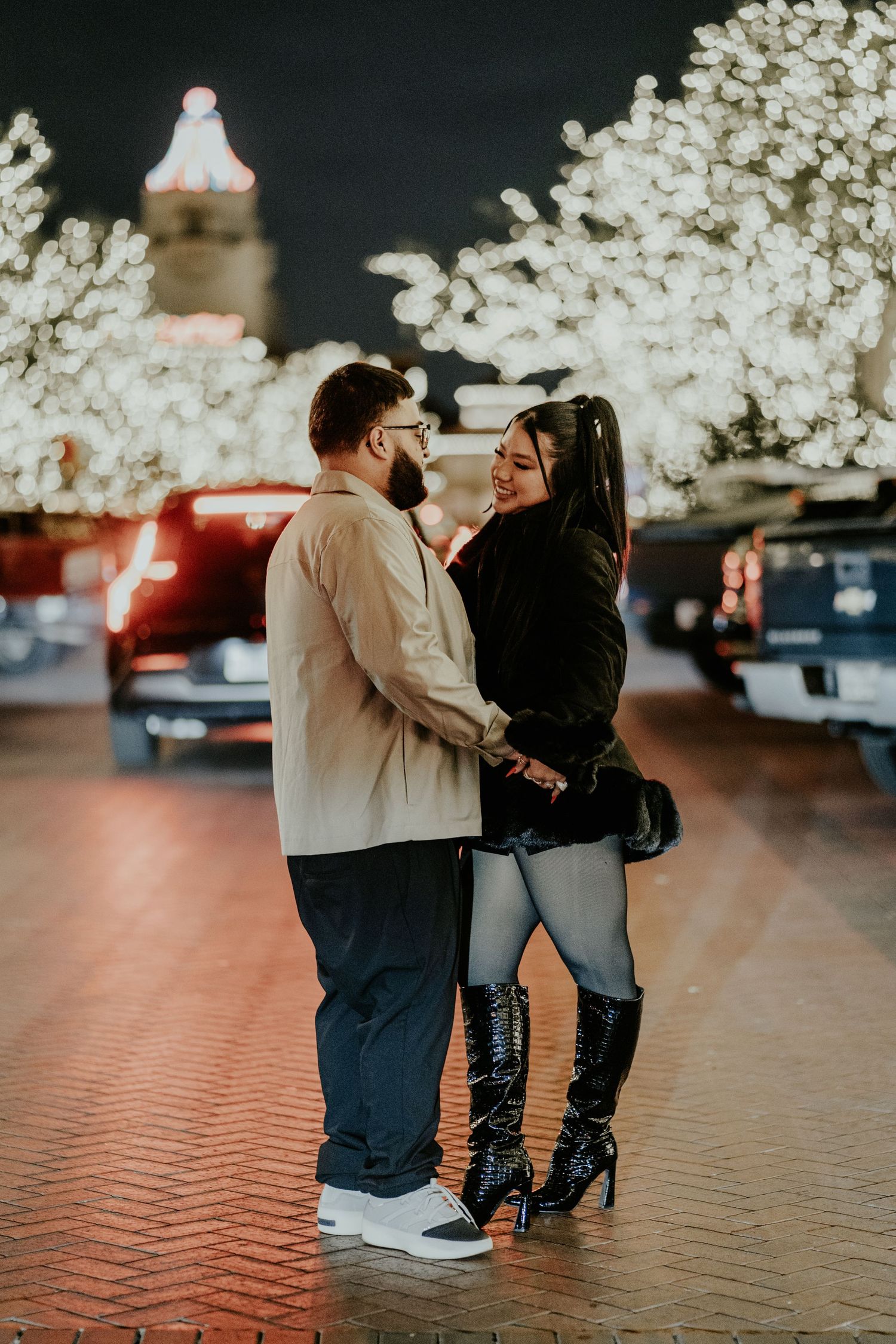 A couple holds hands and smiles under glowing white holiday trees on a brick plaza at night.