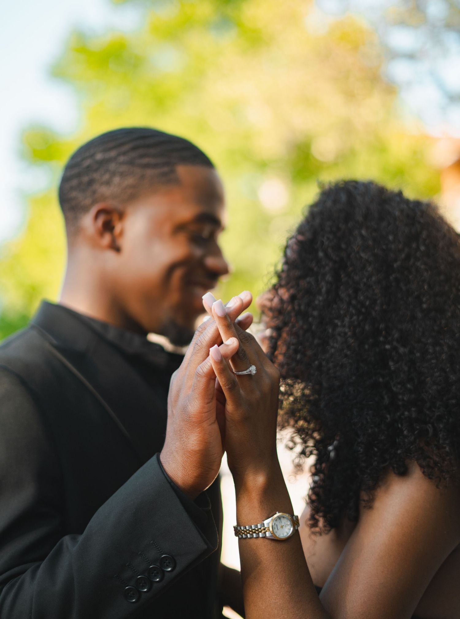 Close-up of a Black couple holding hands and smiling tenderly, with a sparkling engagement ring visible.