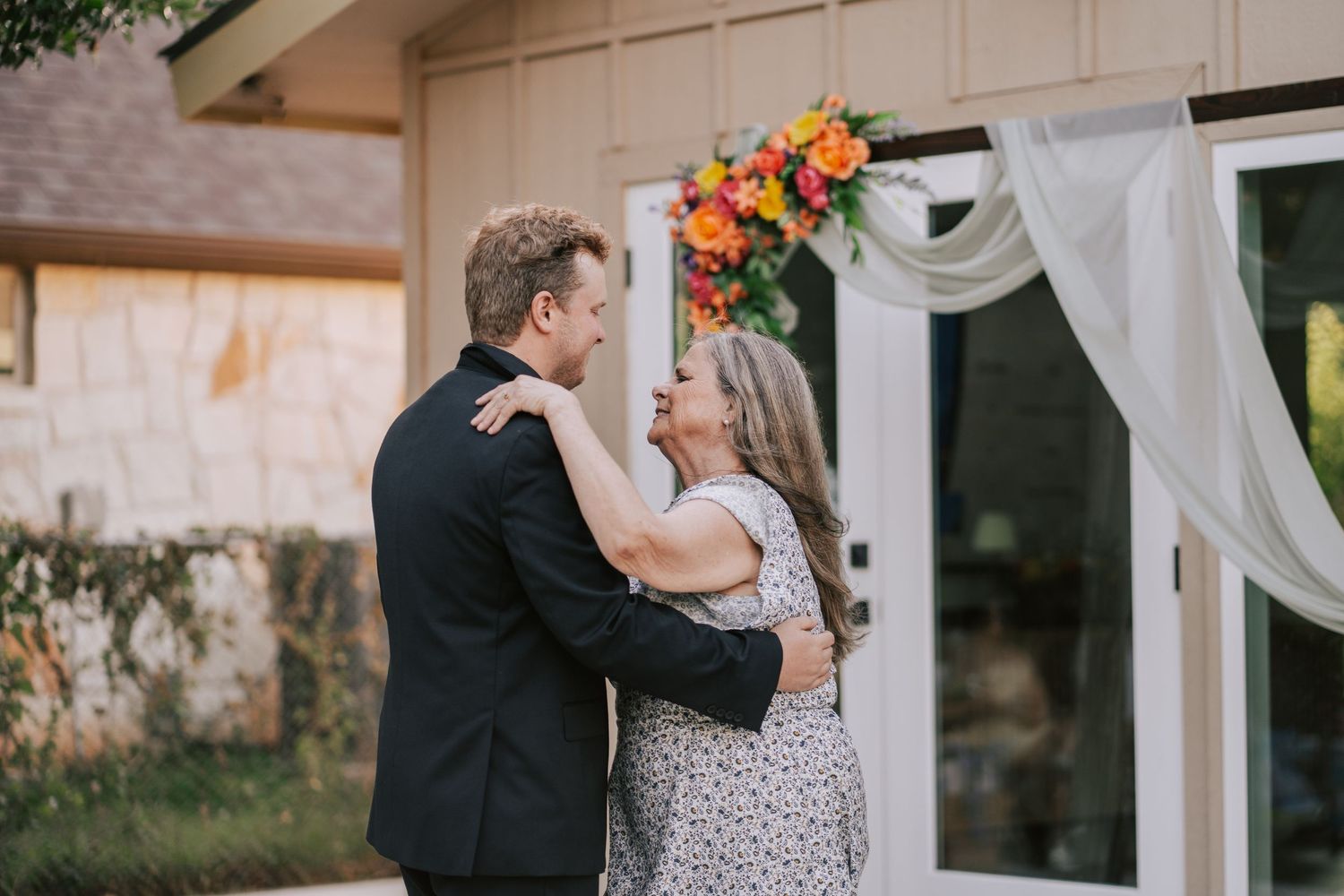 A groom in a black suit embraces a woman in a floral dress outdoors near a colorful flower arrangement at a wedding.