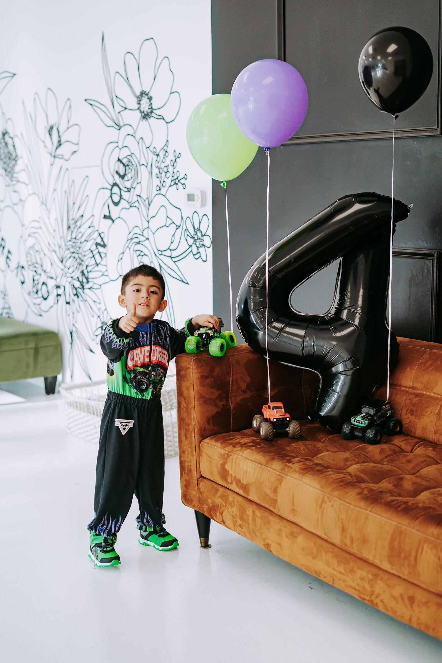 Boy in Monster Jam costume holds toy trucks beside number 4 balloon at birthday party celebration.