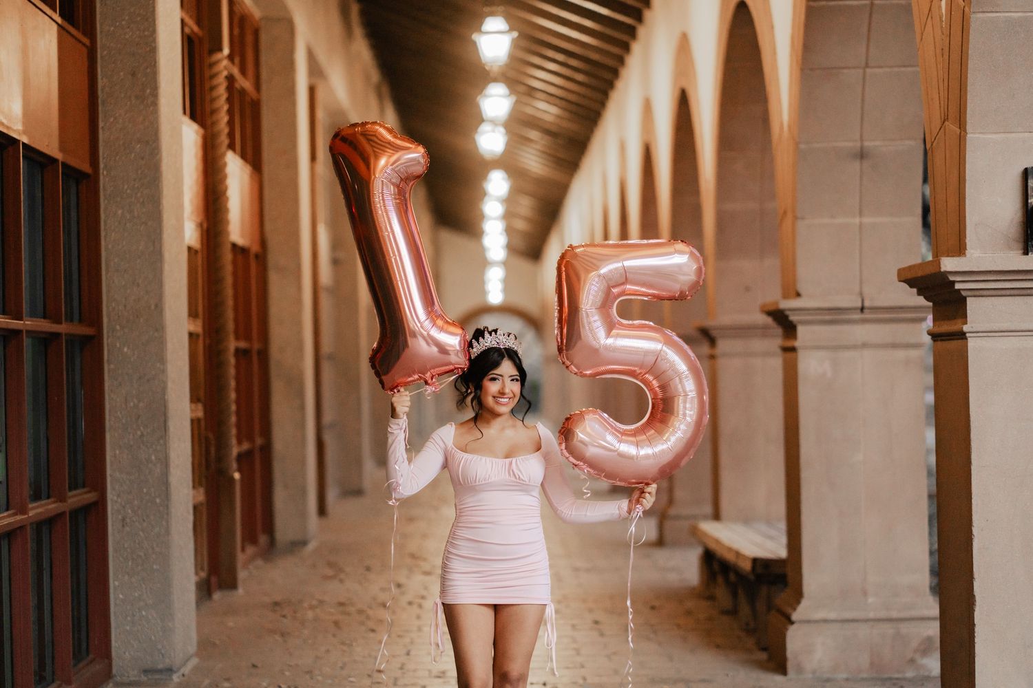 Smiling young woman holds rose gold number 15 balloons in an elegant corridor for her quinceañera.