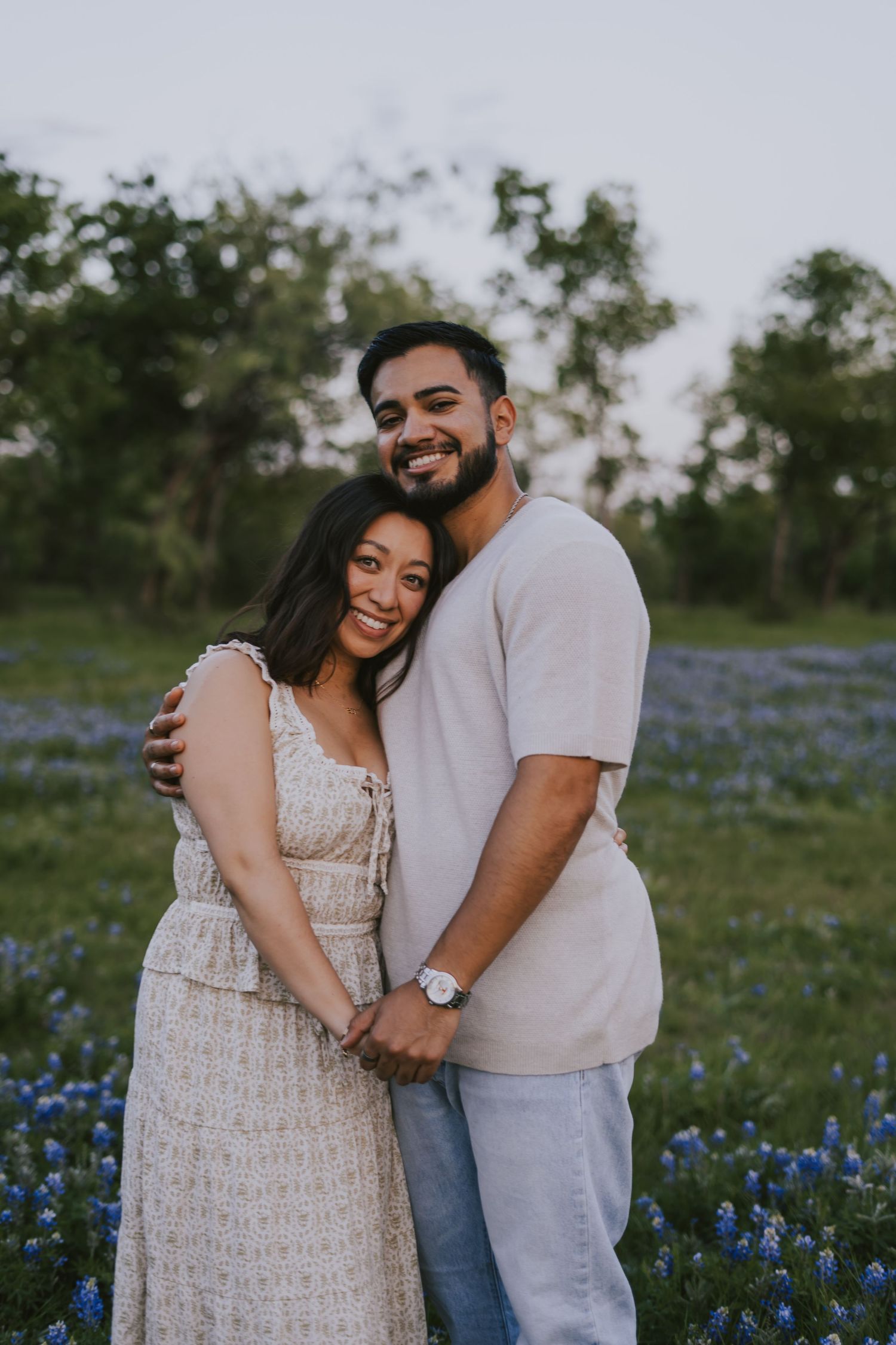 A smiling couple embraces in a field of blue wildflowers, wearing casual white outfits in a lush green outdoor setting.