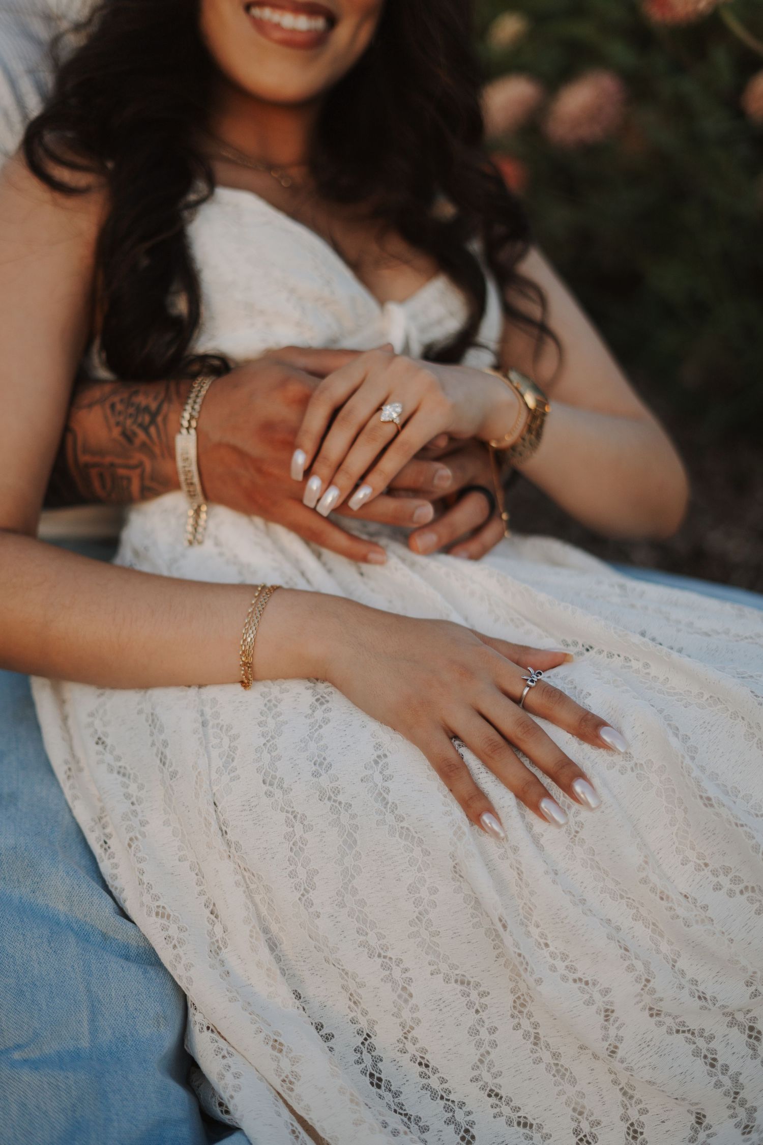 Close-up of an engaged couple holding hands, showcasing a diamond ring, gold bracelets, and white manicure.