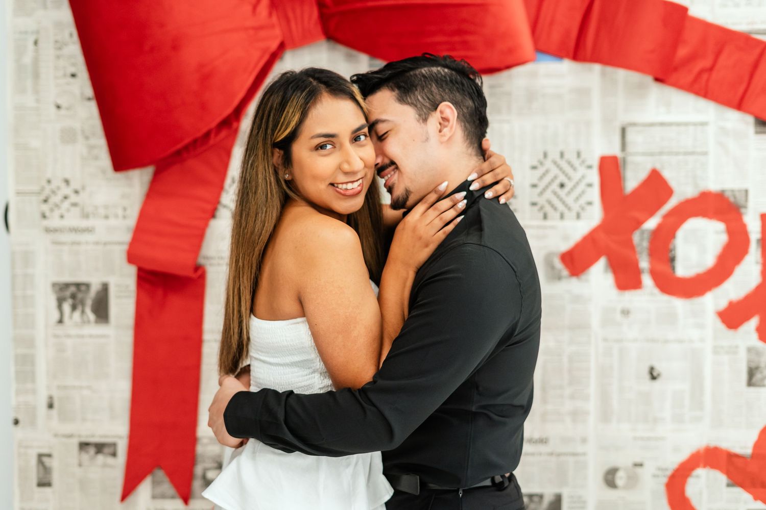 Couple embracing and smiling in front of Valentine's Day XOXO newspaper backdrop with red bow.