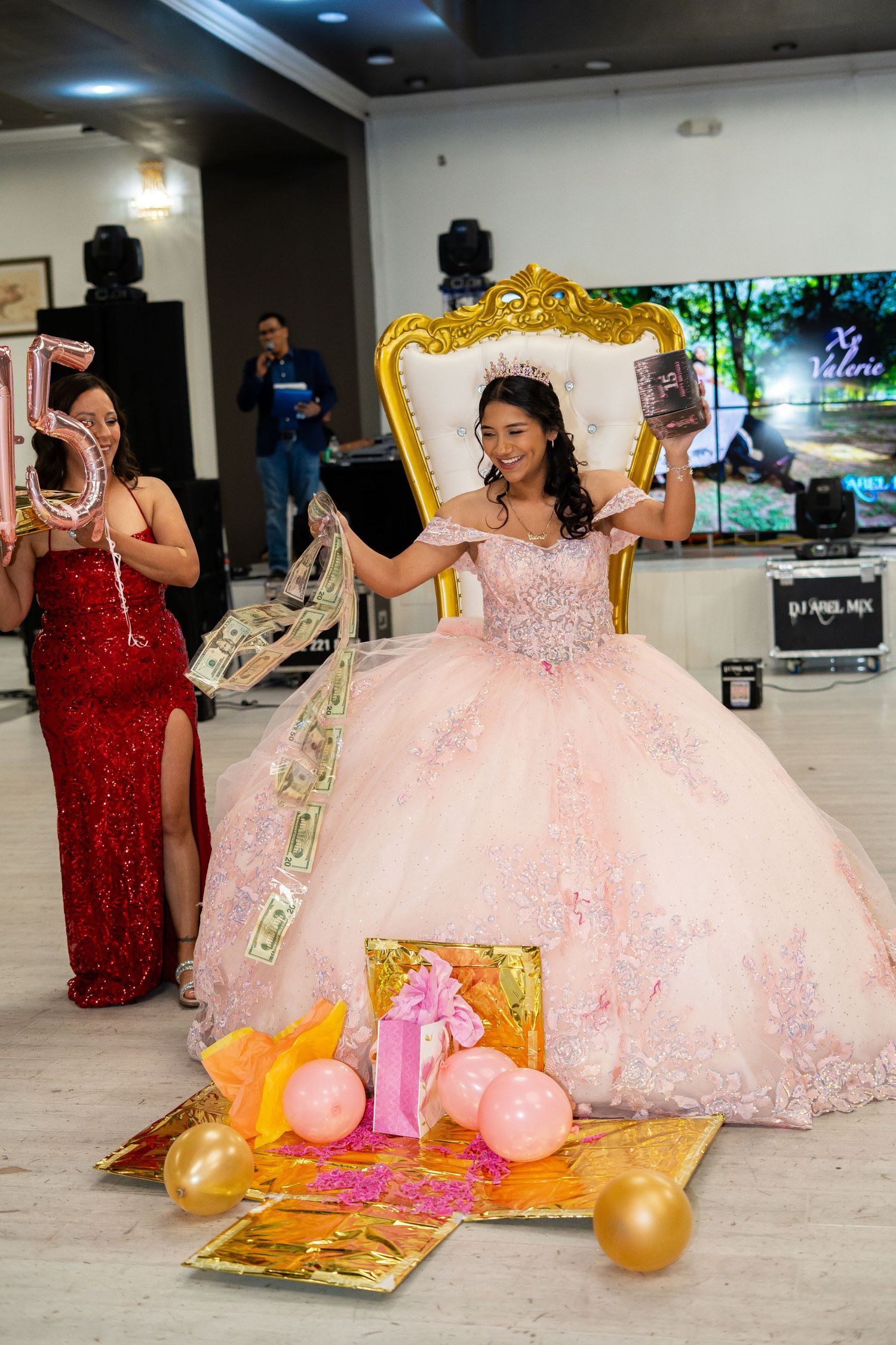 A quinceañera in a pink gown opens gifts while seated on a gold throne.