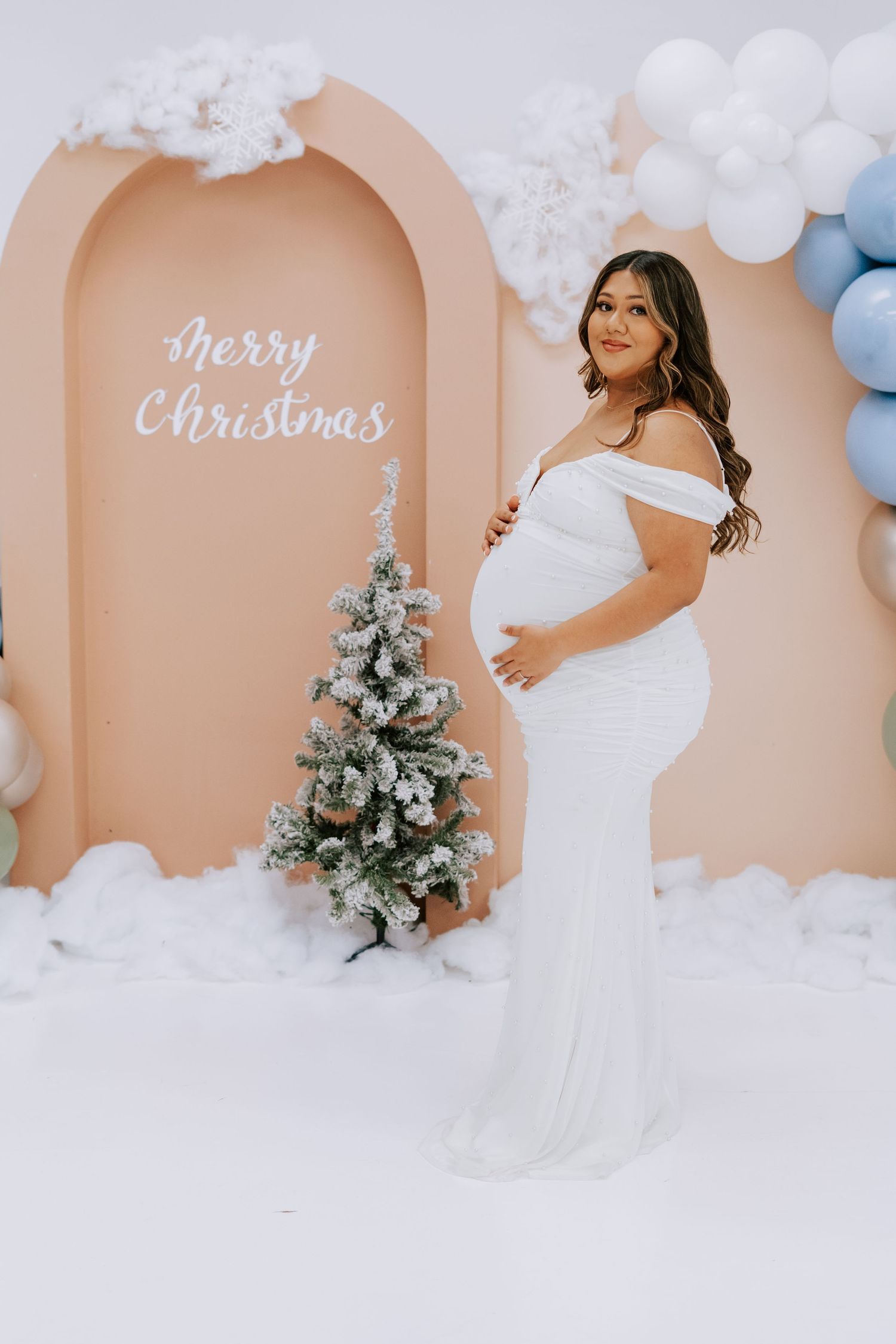 Pregnant woman in white off-shoulder gown posing by Christmas tree with Merry Christmas backdrop.