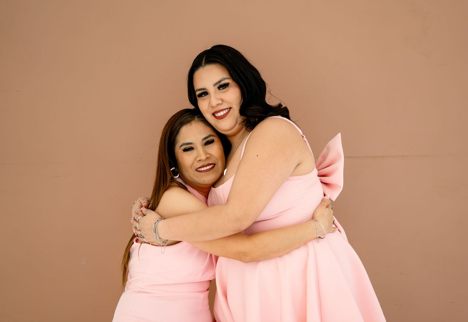 Two women in pink dresses share a warm embrace in a studio portrait.