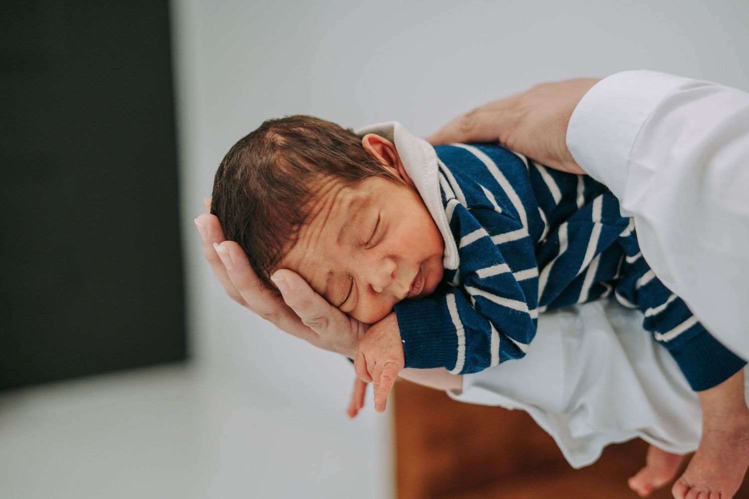 A newborn baby in a blue striped outfit resting peacefully in a parent's supportive hands.
