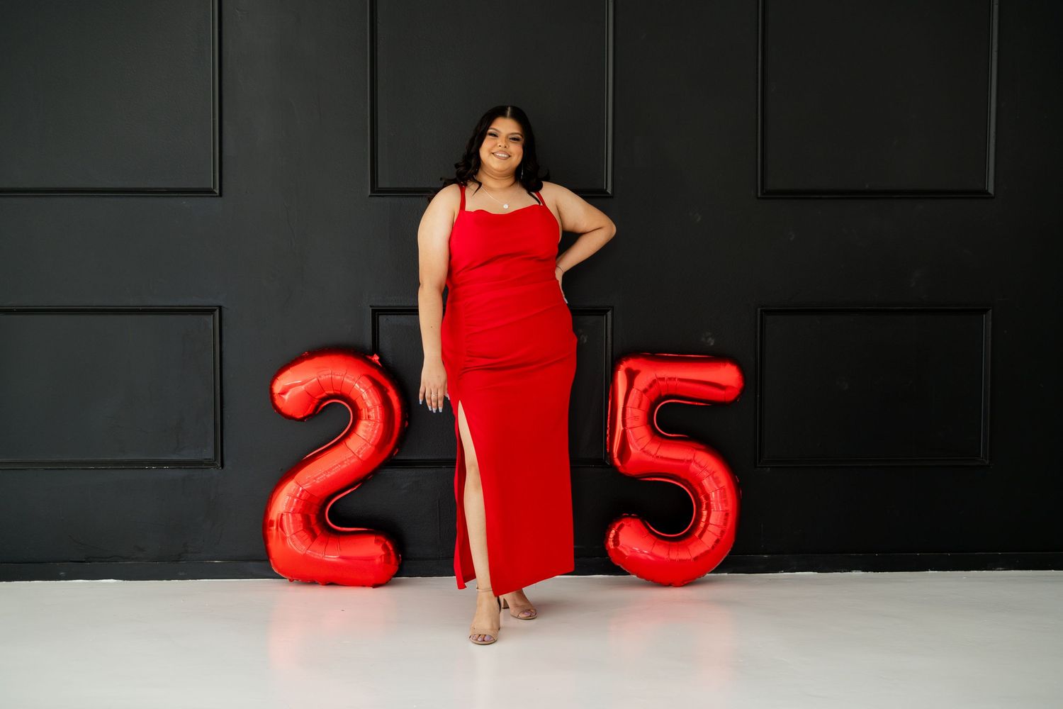 A woman in a red dress stands confidently beside large red number 25 balloons.