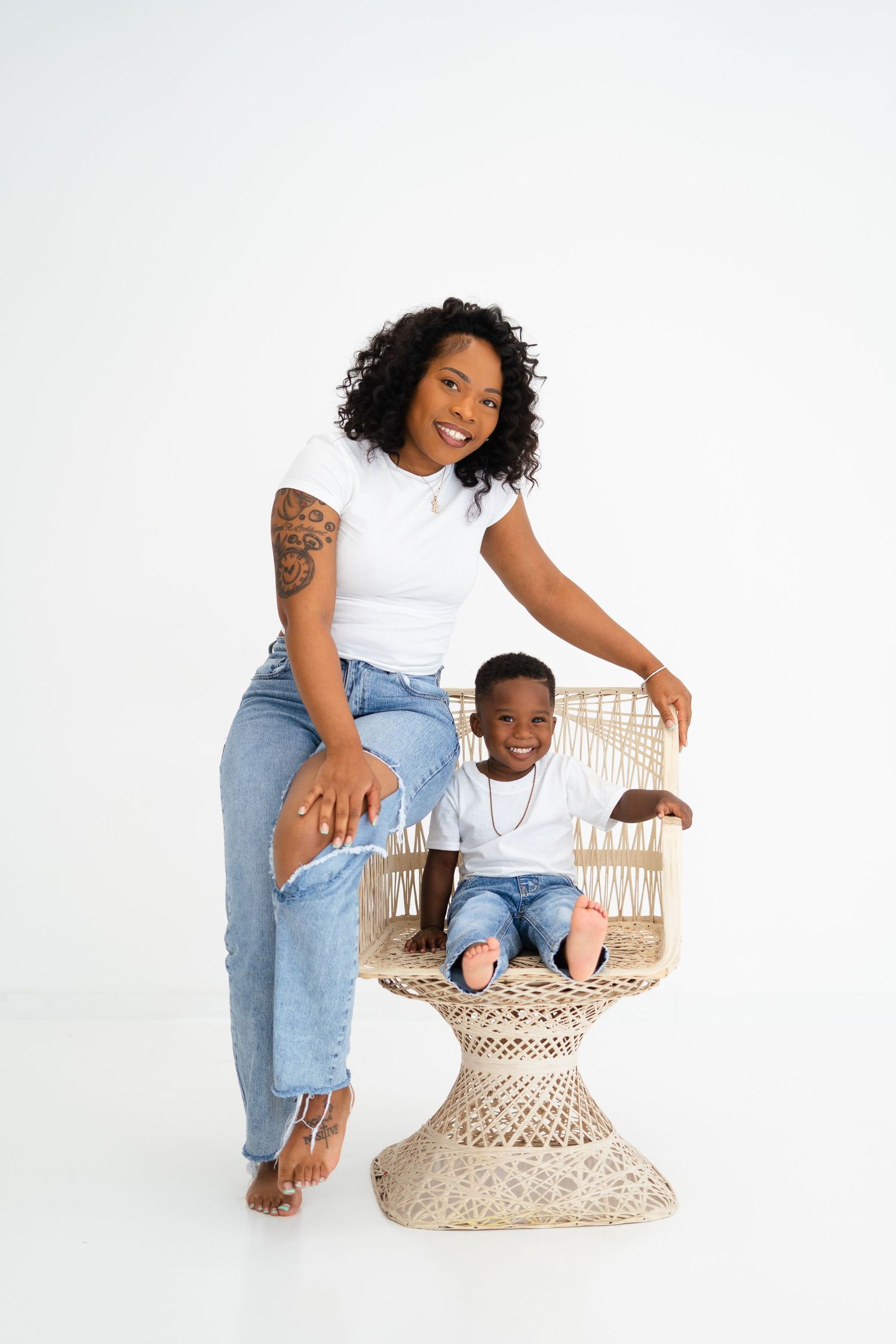 Smiling Black mother perched on wicker chair beside her toddler son in white studio photo.