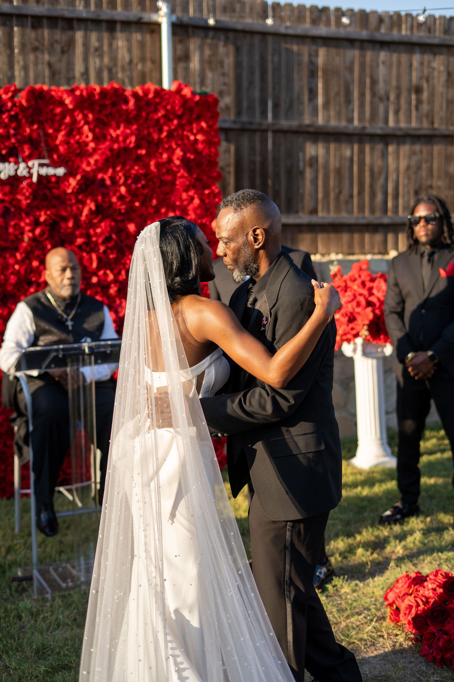 Newlyweds share first kiss at outdoor wedding ceremony in front of red rose flower wall backdrop.