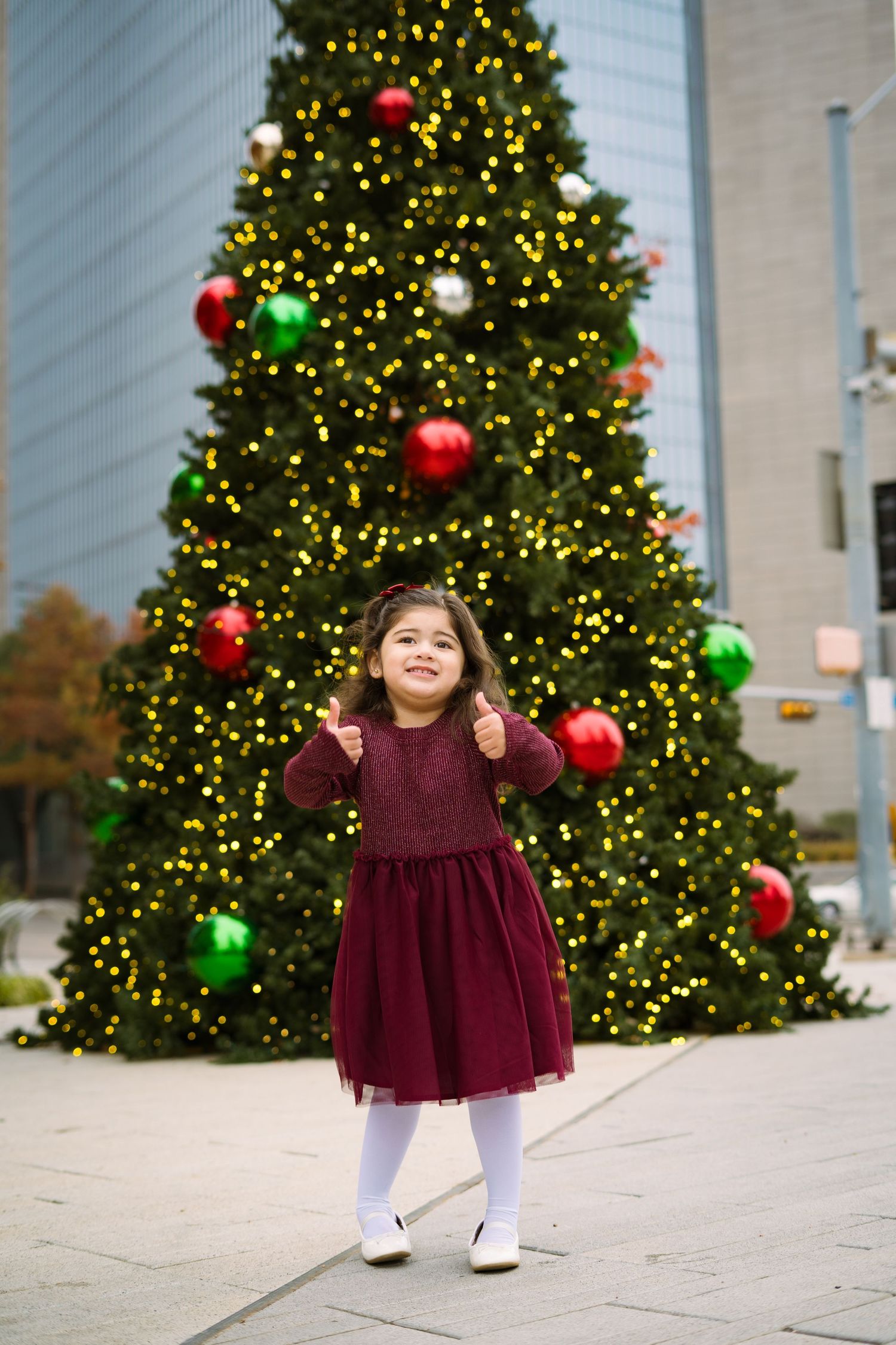 A joyful little girl in a burgundy dress gives thumbs up in front of a large decorated Christmas tree.