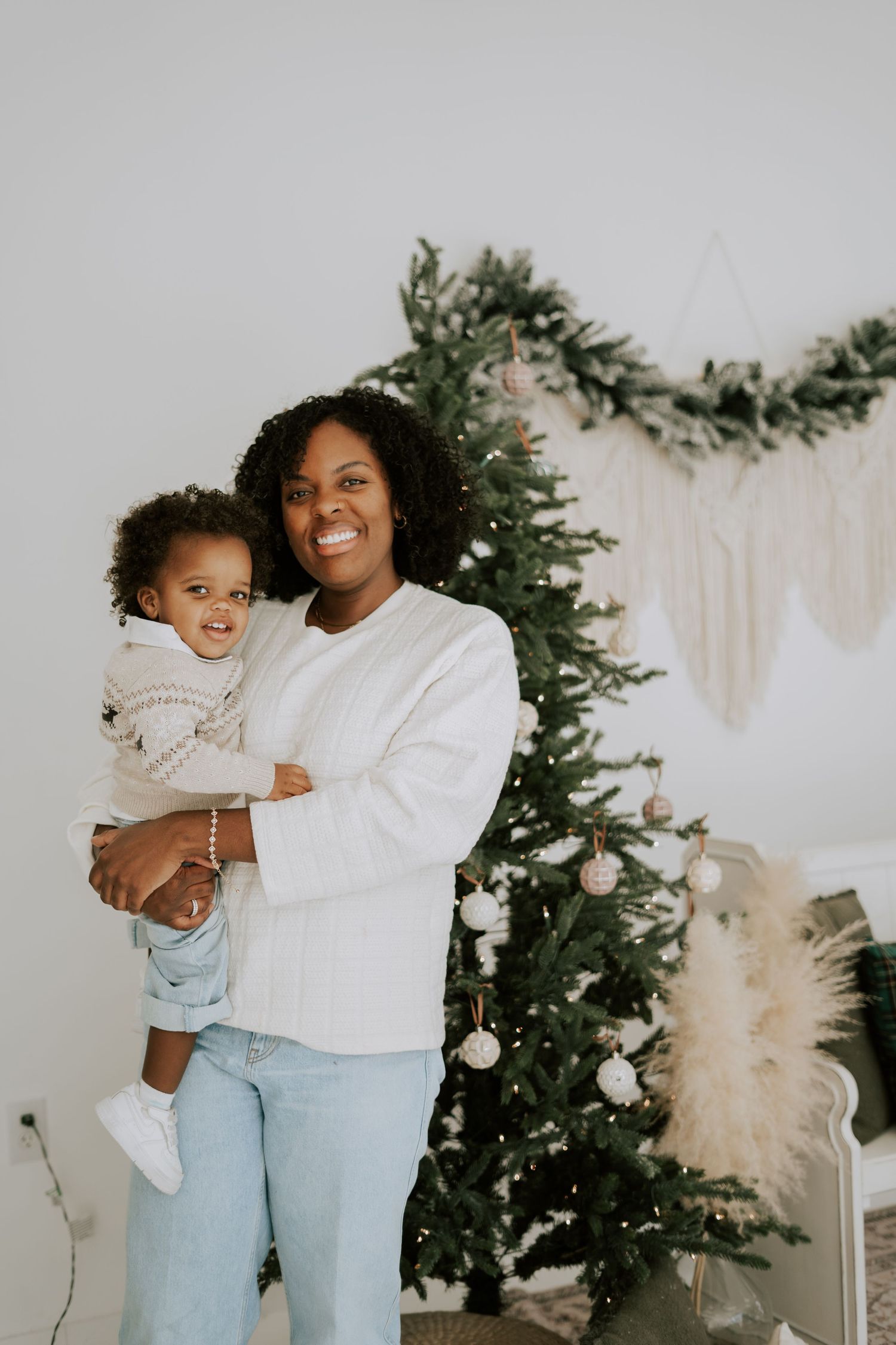 Happy mother in white sweater holds smiling toddler beside decorated holiday Christmas tree.