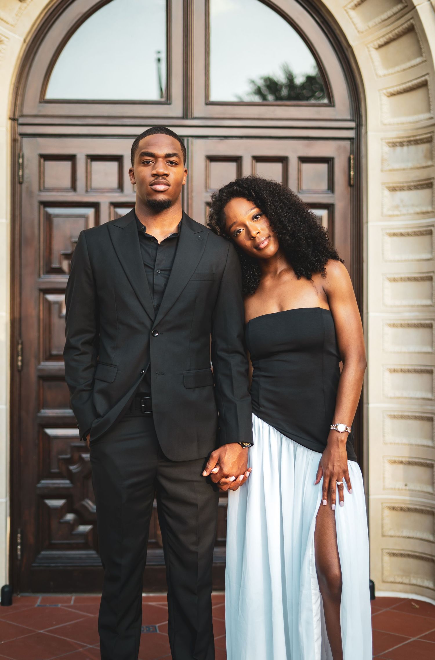 Black couple holding hands in formal black and white attire before ornate wooden doors.