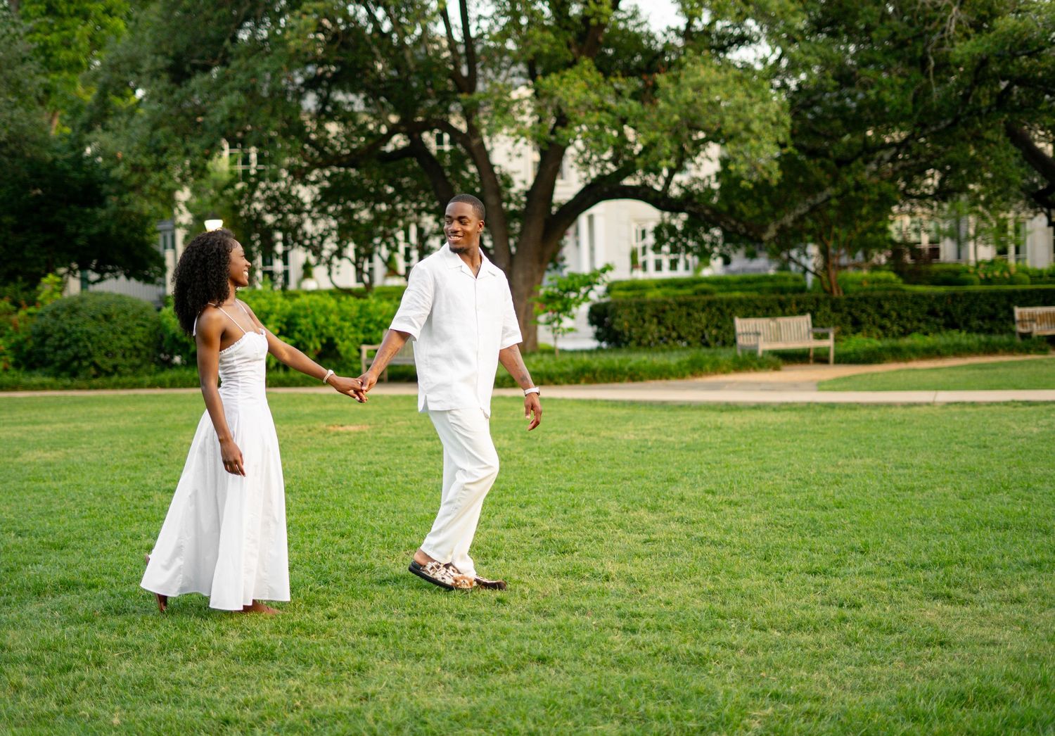 A joyful Black couple in matching white outfits holds hands while walking on lush green grass in a sunny park.