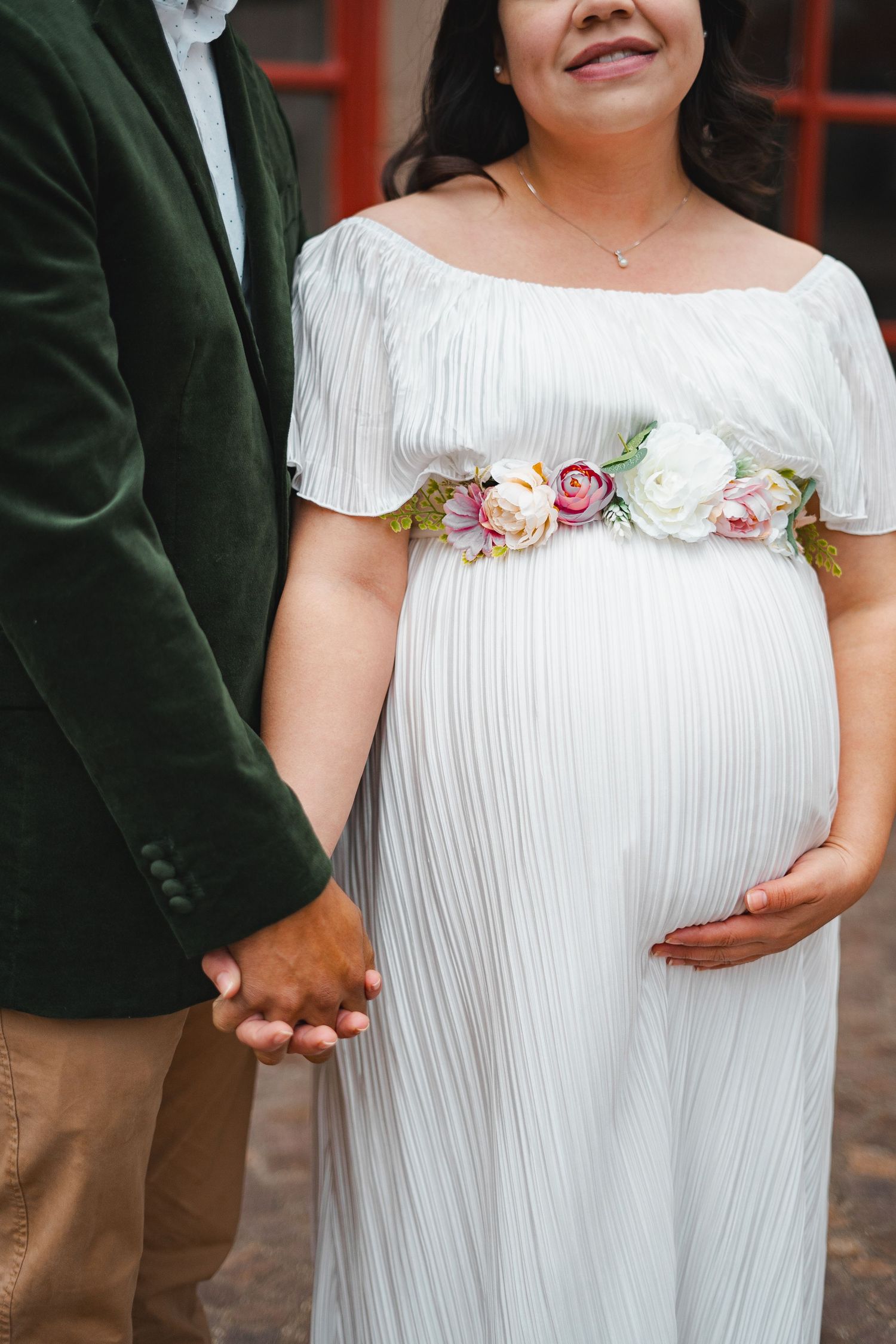 Pregnant woman in white off-shoulder maternity dress with floral sash holding hands with partner.