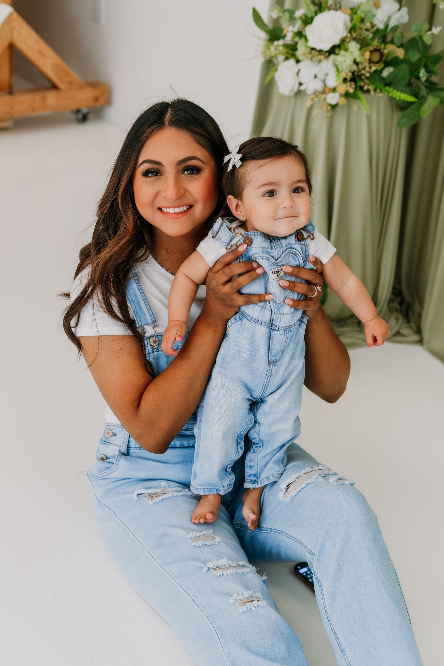 A young woman and baby in matching denim overalls pose together for a studio photo session.