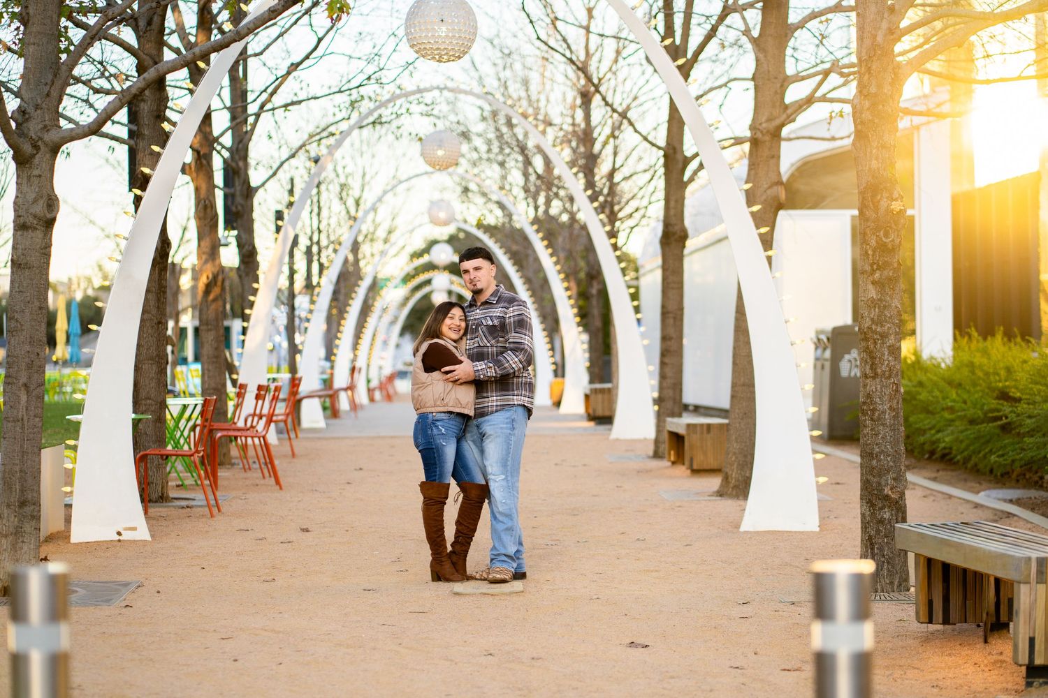 A couple embraces warmly on a park pathway surrounded by bare trees and glowing sunset light.