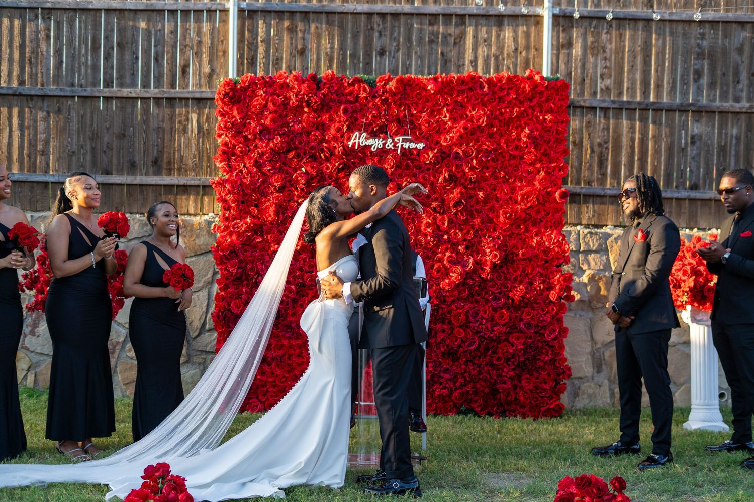 Bride and groom share first kiss in front of red rose flower wall surrounded by wedding party.