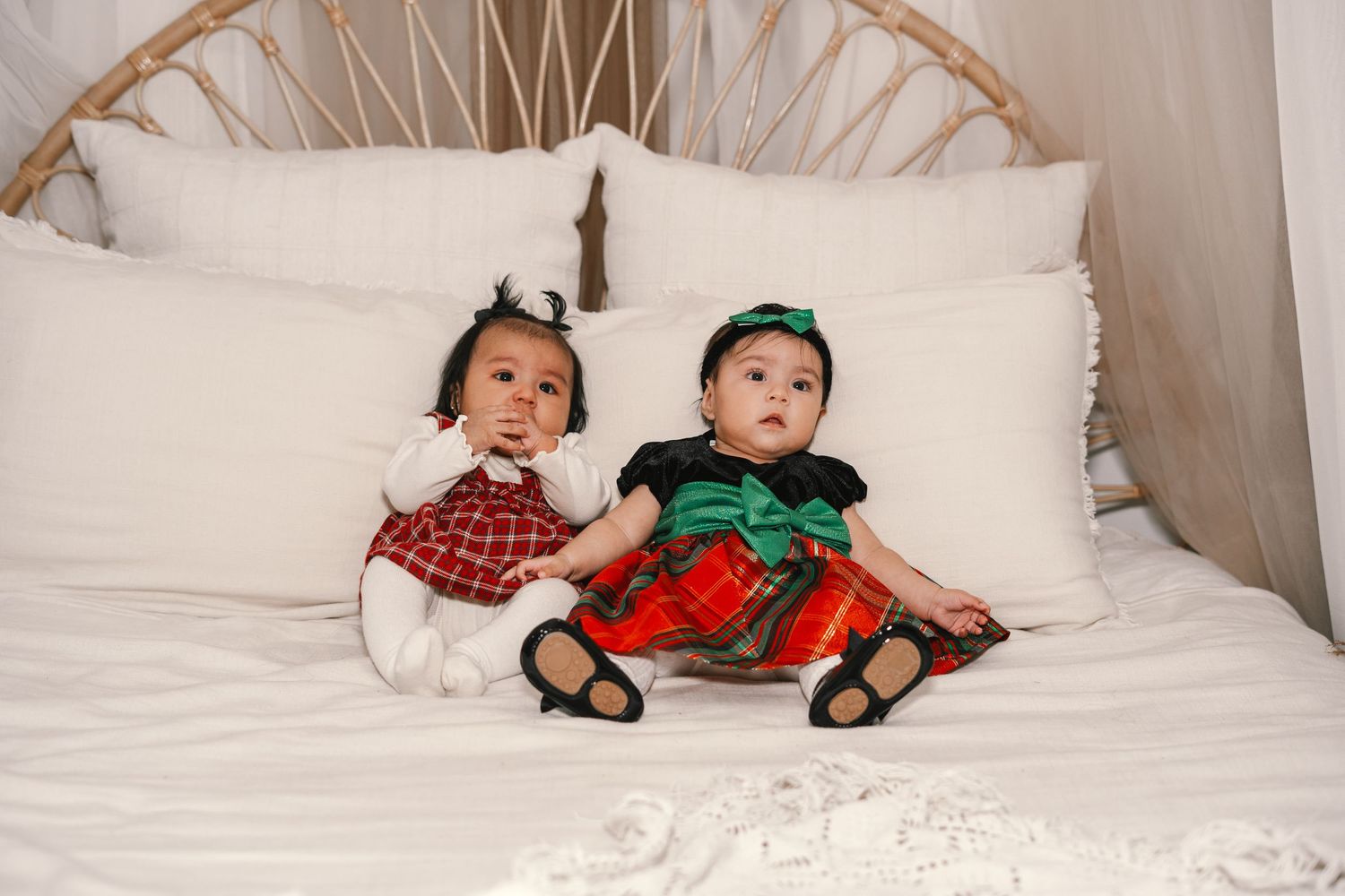 Two baby girls in red and green Christmas dresses sit together on a white bed with pillows.