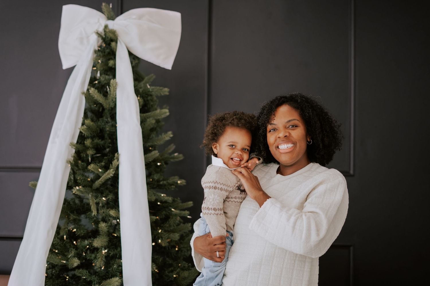 A joyful mother holds her smiling baby boy next to a Christmas tree adorned with a white ribbon bow.
