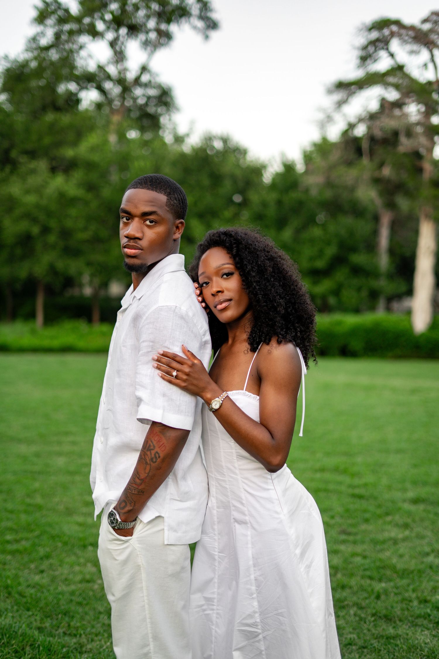 A Black couple in white poses back-to-back on green grass, with the woman embracing the man from behind.