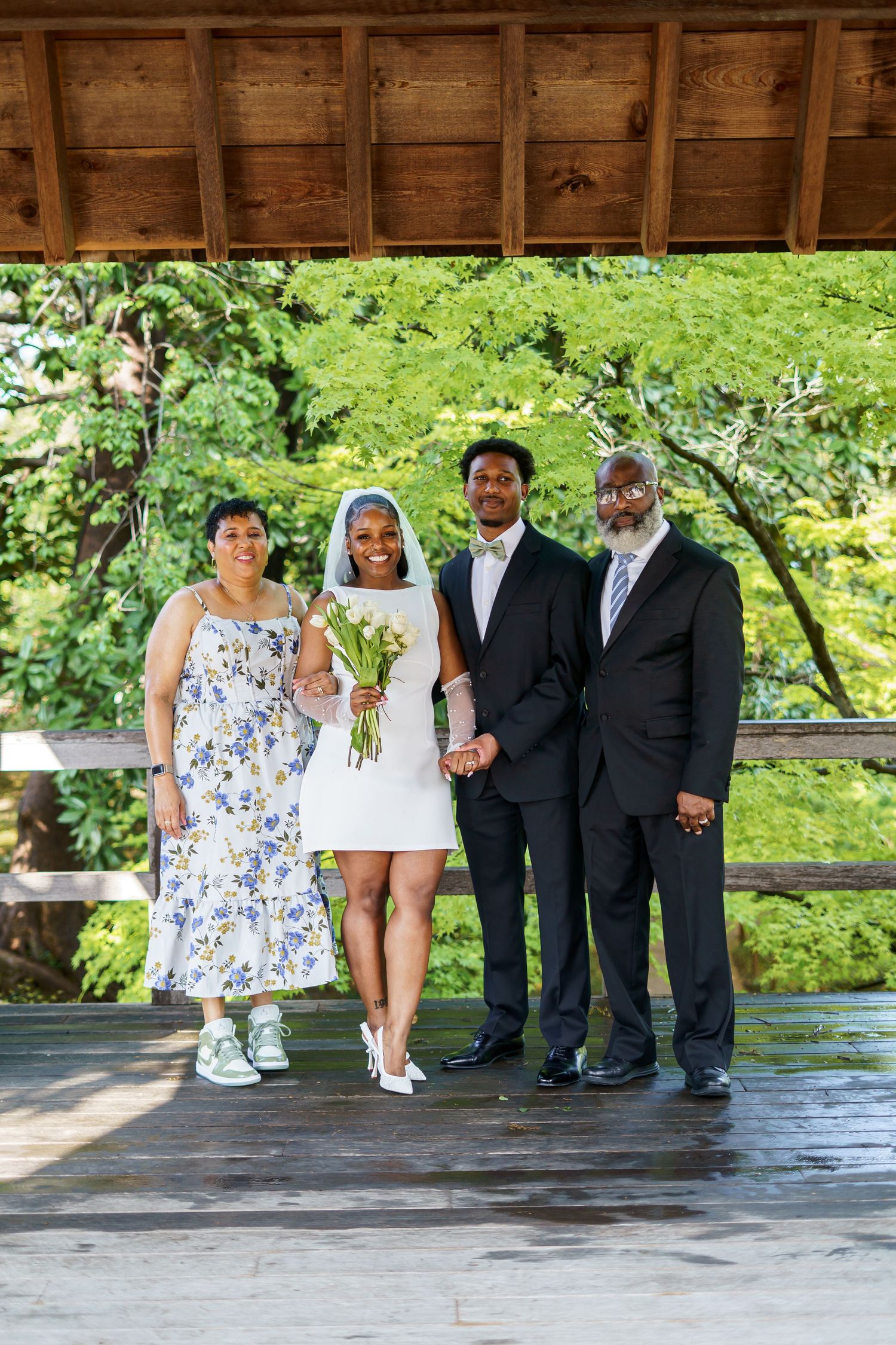 Bride holding white tulip bouquet poses with groom and two family members under a wooden pavilion.