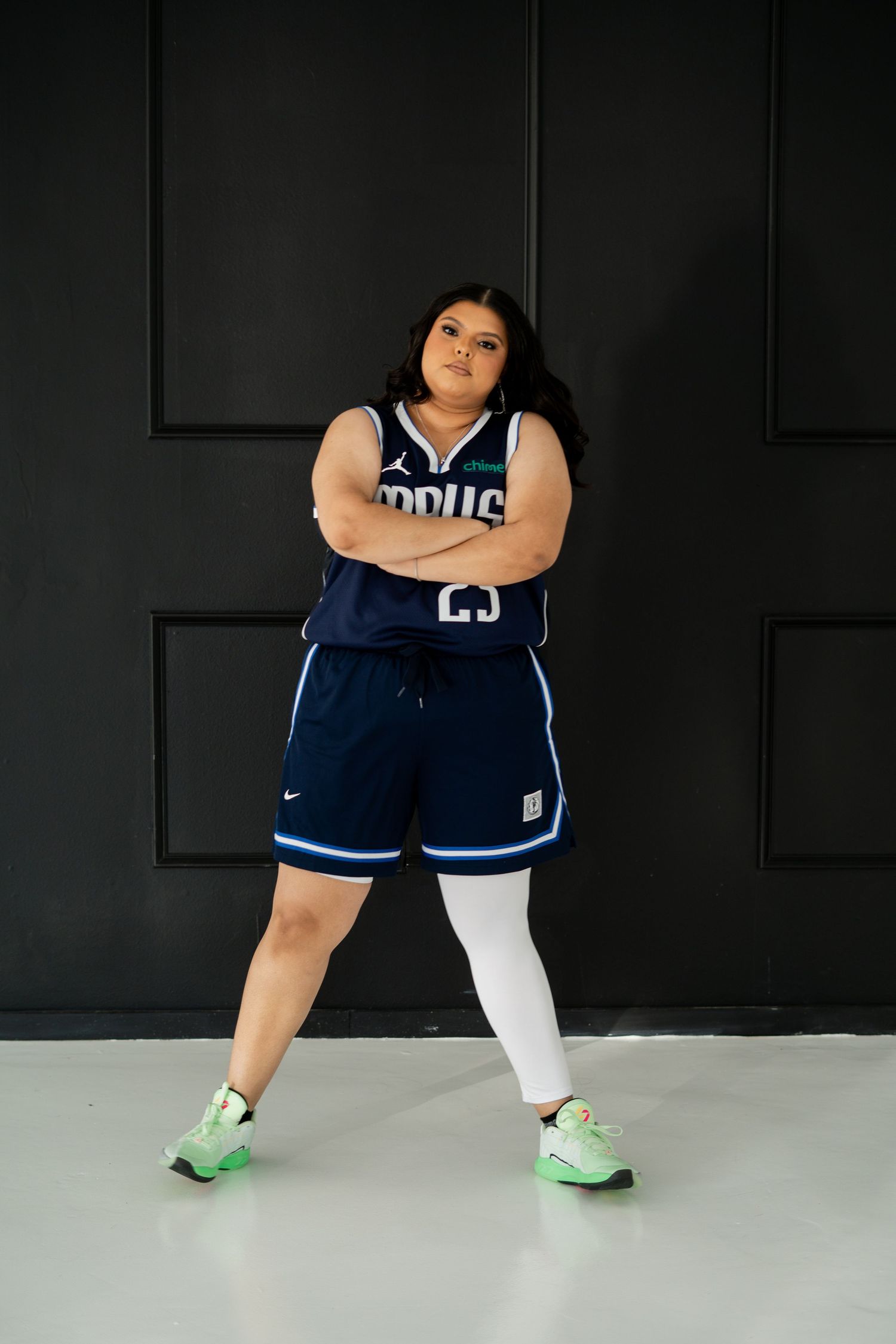 Woman in Dallas Mavericks jersey and shorts posing with arms crossed against black wall.