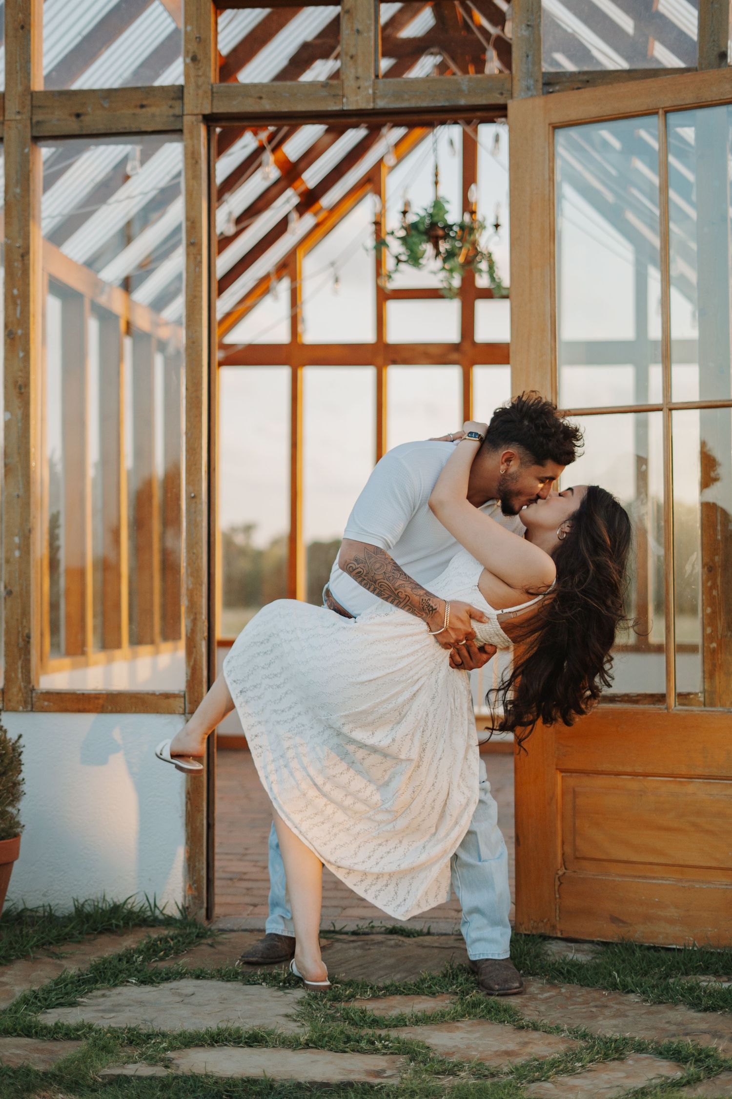 Couple sharing a romantic dip kiss in front of a rustic greenhouse at sunset, woman in white skirt.