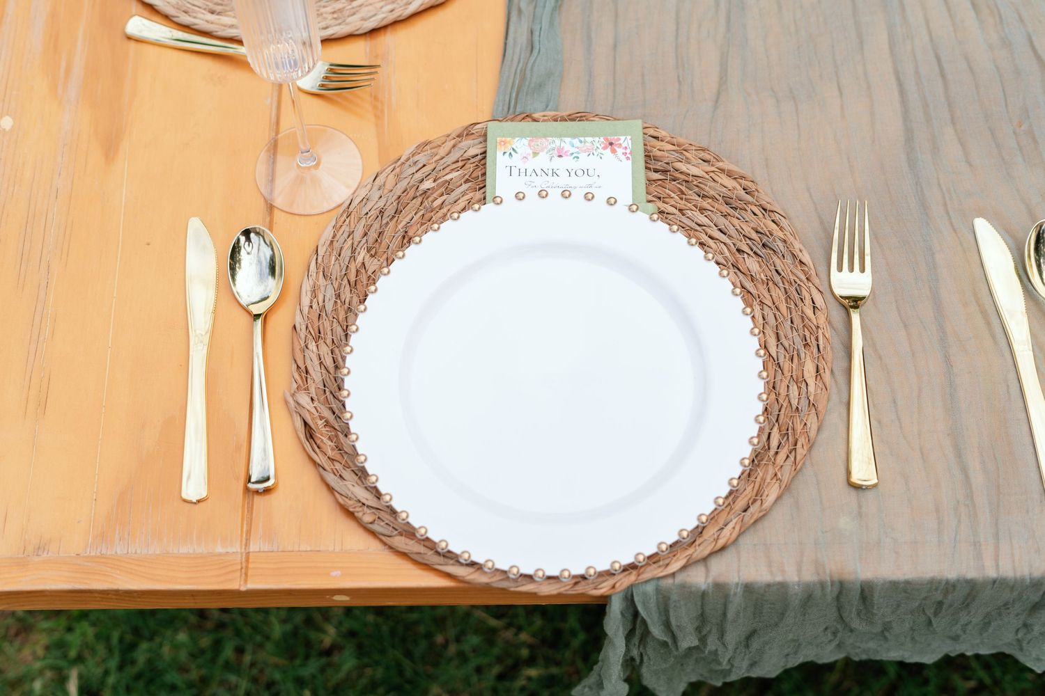 Elegant rustic place setting with white plate, gold cutlery, woven charger, and floral thank you card on wooden table.