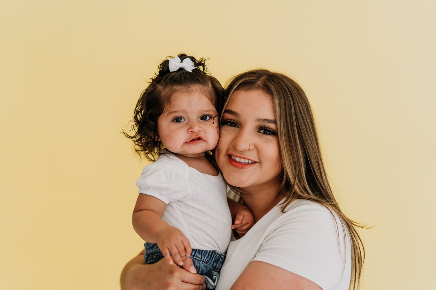 A young woman in a white shirt smiles while holding a cute baby girl with a bow in her dark curly hair.