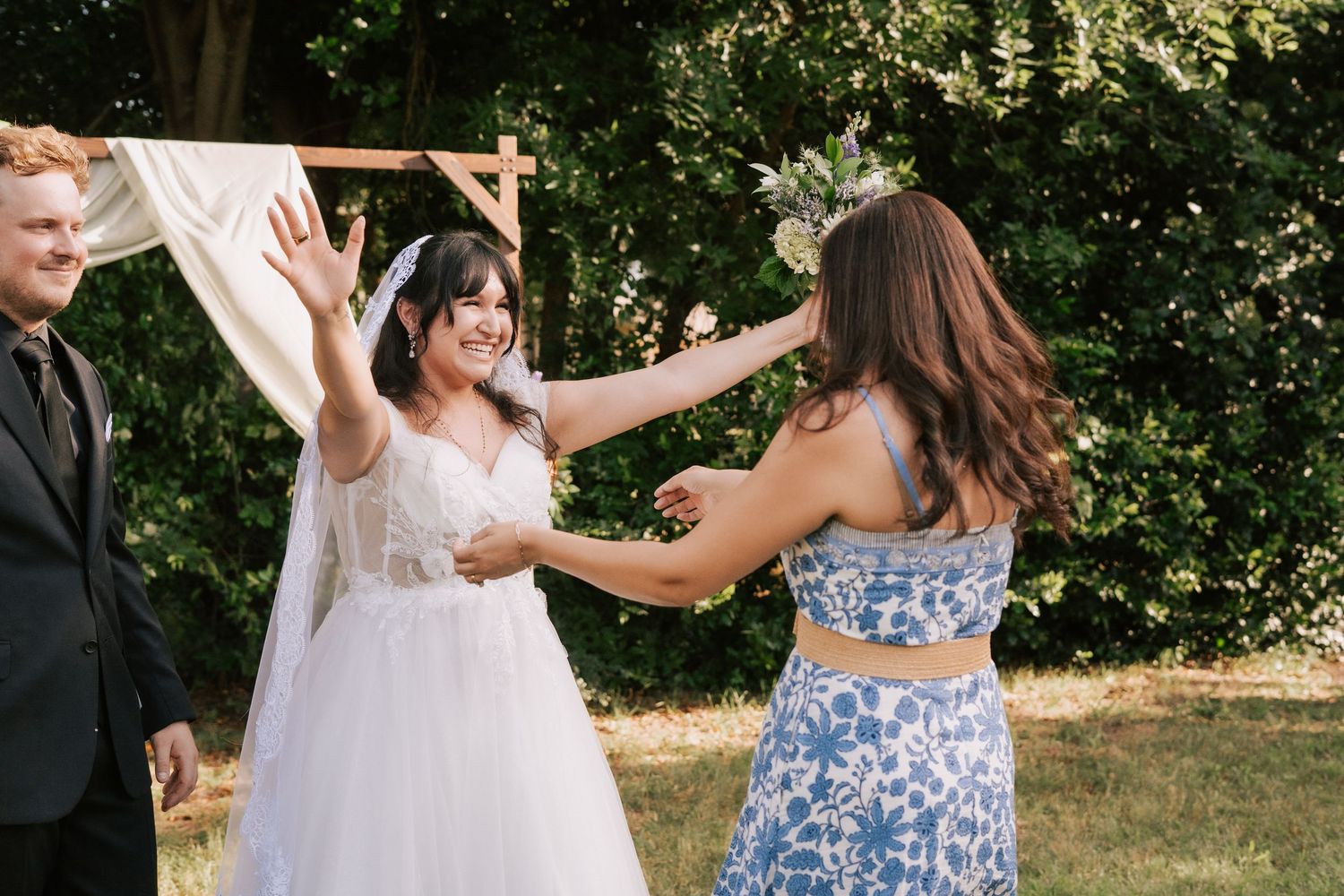 Bride in white gown laughing with bridesmaid in blue floral dress during outdoor wedding ceremony.
