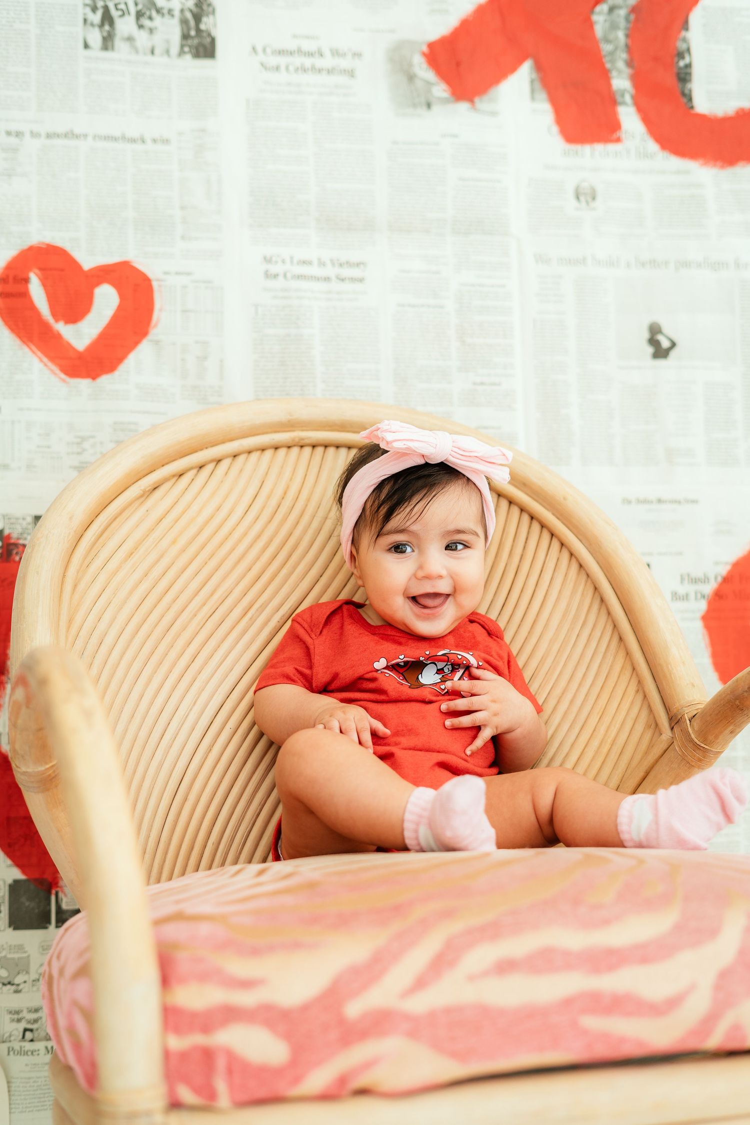 Adorable baby girl in a red outfit and pink bow headband smiling while sitting in a wicker chair.