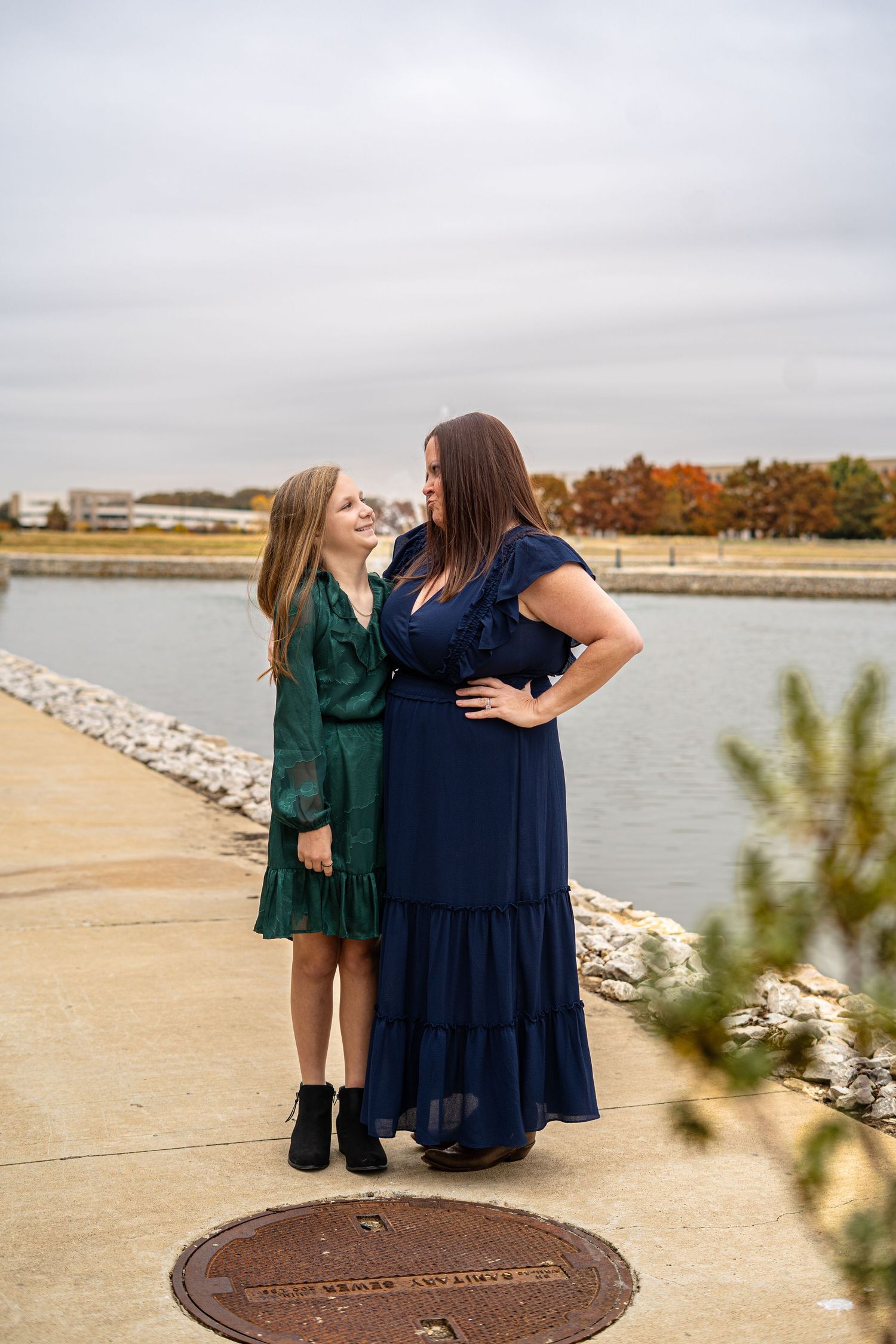 Mother and daughter in blue and green dresses smiling at each other on a lakeside walkway.