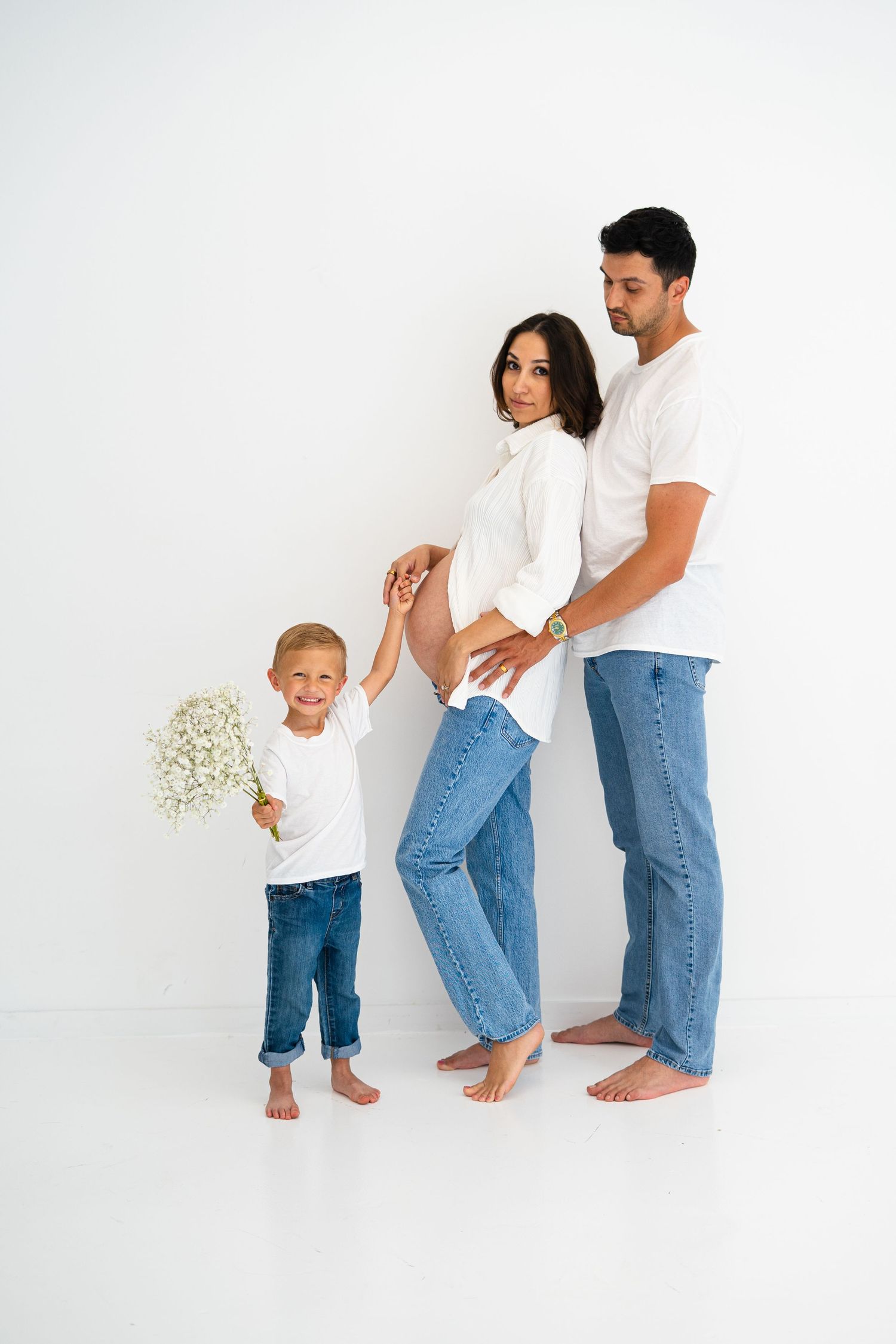 A pregnant woman, her partner, and young son in matching white shirts and jeans pose for a maternity photo.