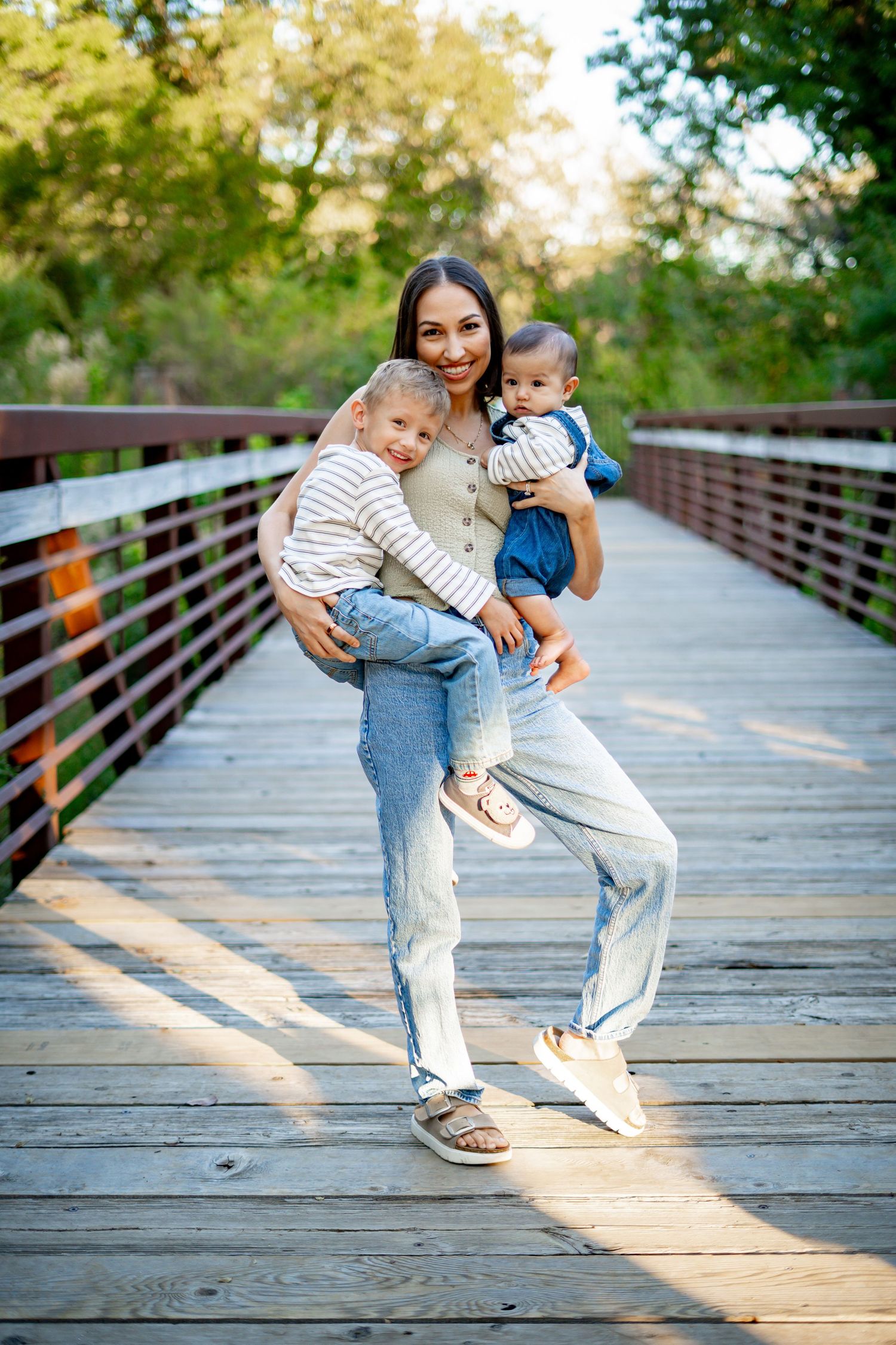 Joyful mother holding two young children on a sunny wooden bridge with green trees behind.