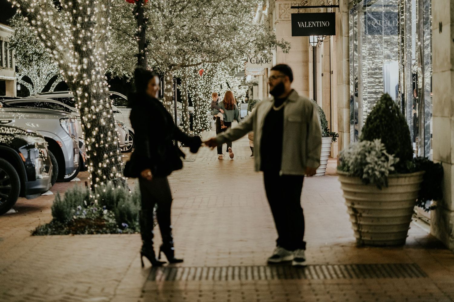 A couple holds hands on a festive lit outdoor shopping promenade at night.