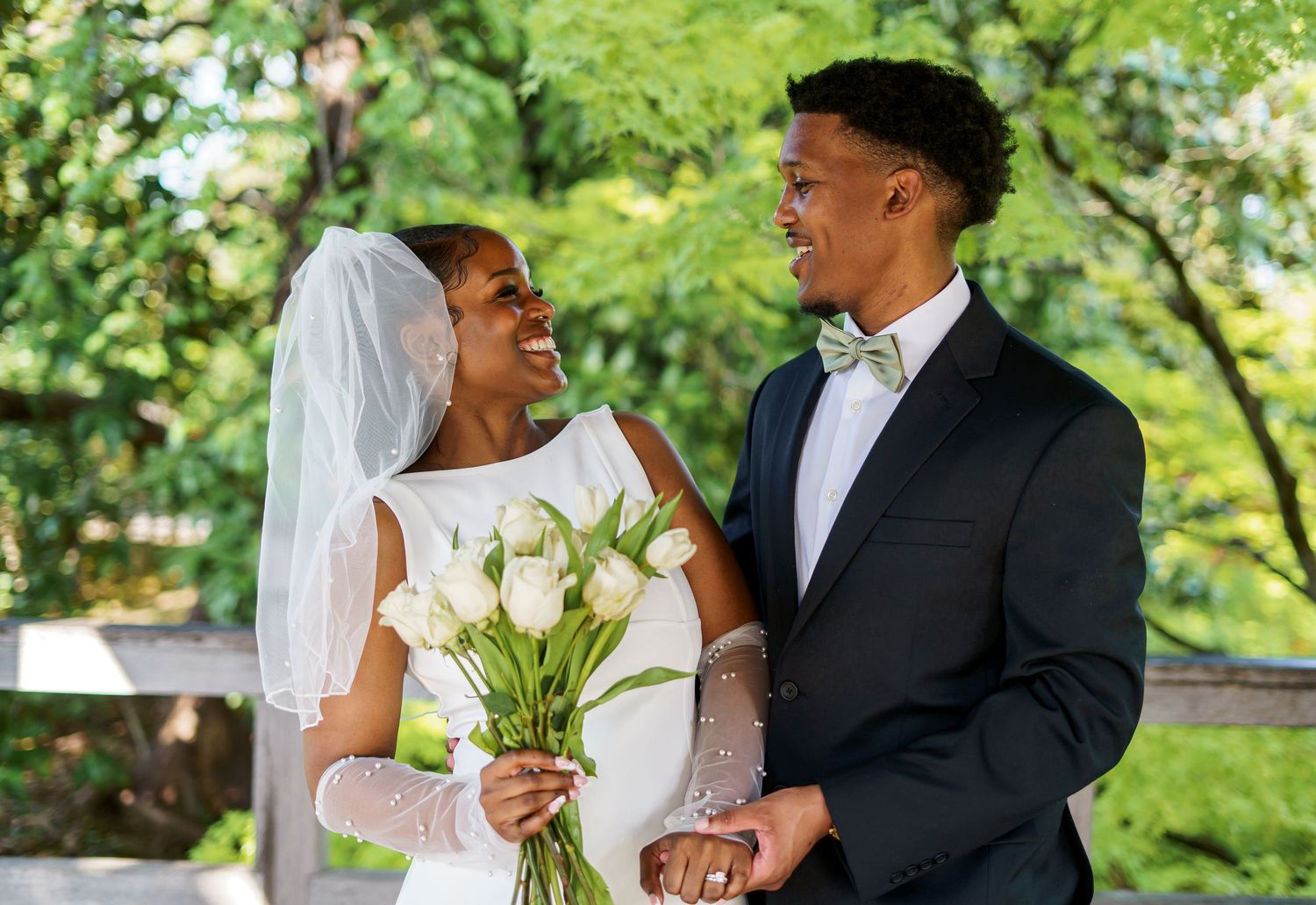 Happy newlyweds share a joyful moment outdoors, bride holding white tulips in wedding dress and veil, groom in navy suit.