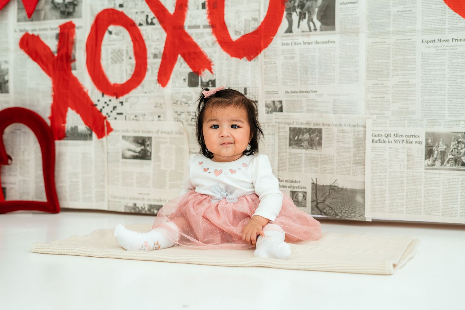 Baby girl in pink skirt and white top sits on mat before XOXO Valentine's Day backdrop.