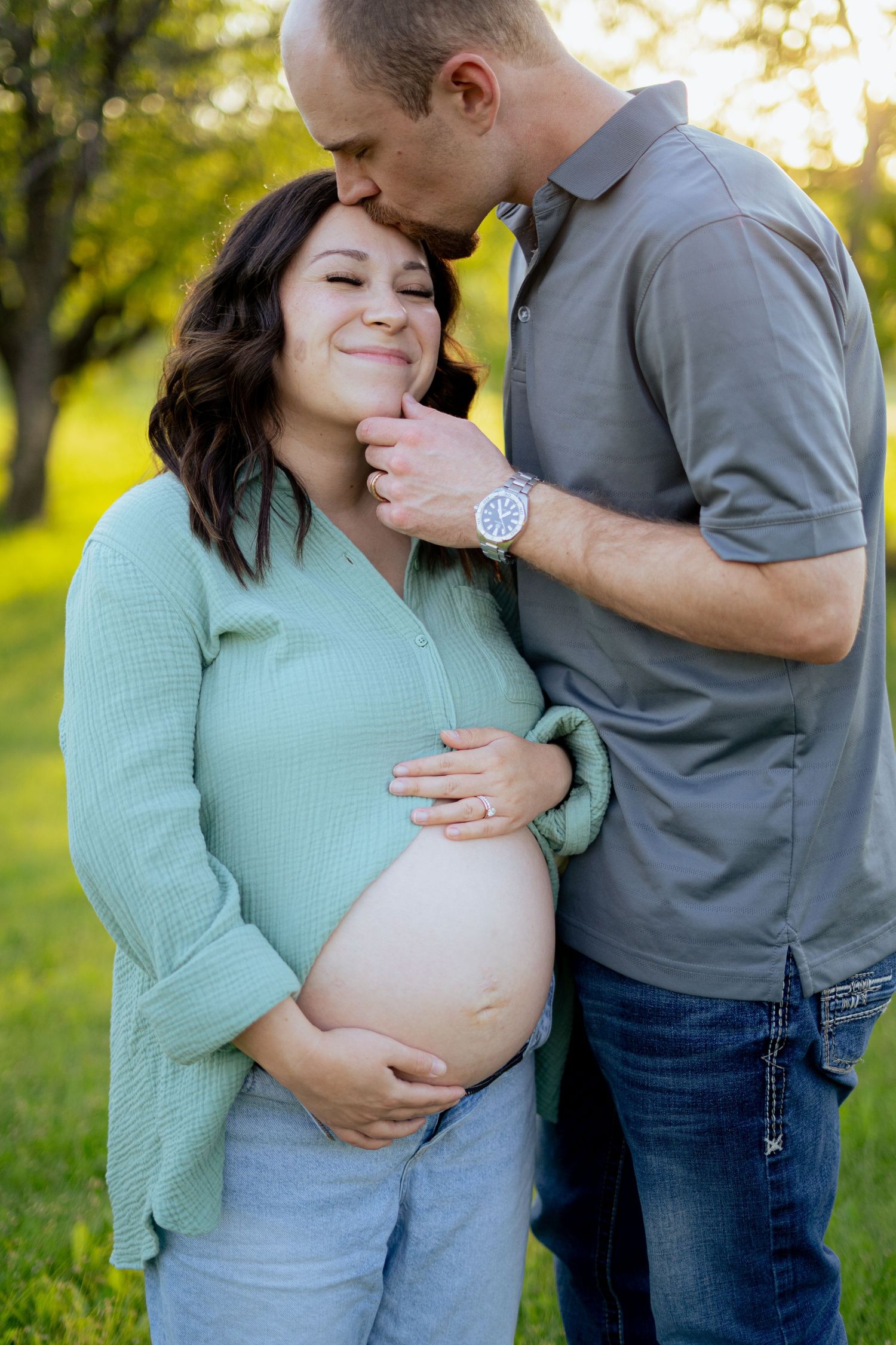 Man tenderly kisses pregnant woman on cheek while gently holding her baby bump in sunny outdoor maternity session.