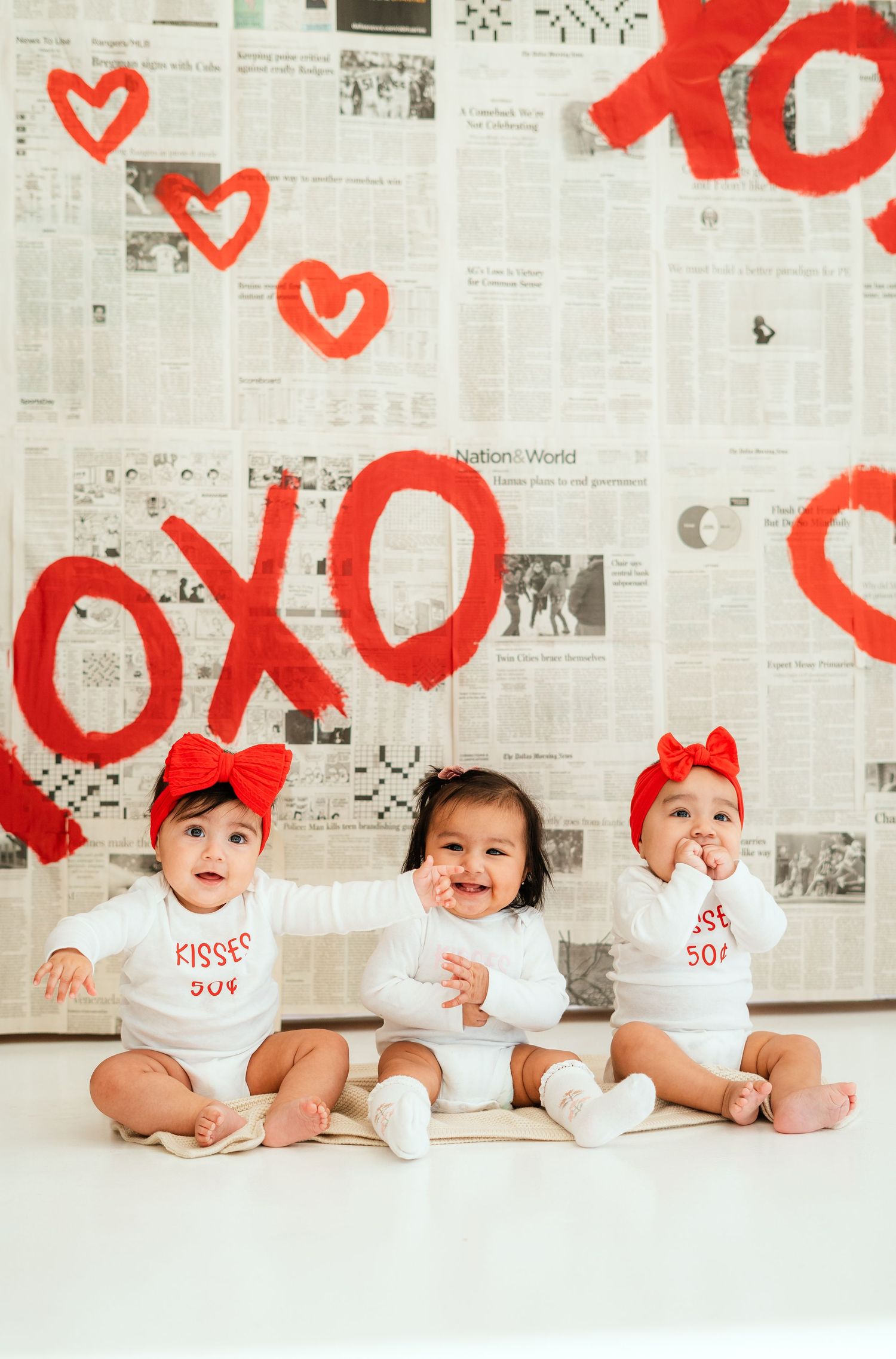 Three babies in white Valentine onesies with red bows sit before a newspaper backdrop painted with red XOXO hearts.
