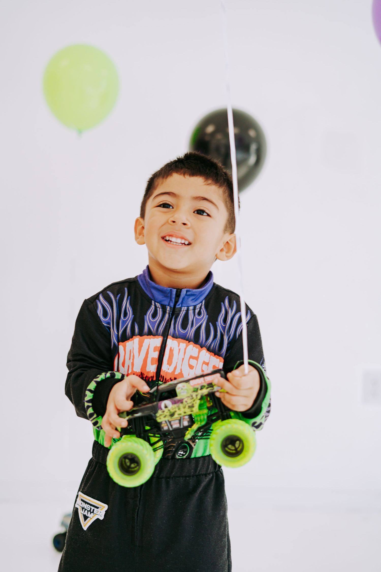 Smiling boy holding Grave Digger monster truck toy with green and black balloons behind him.