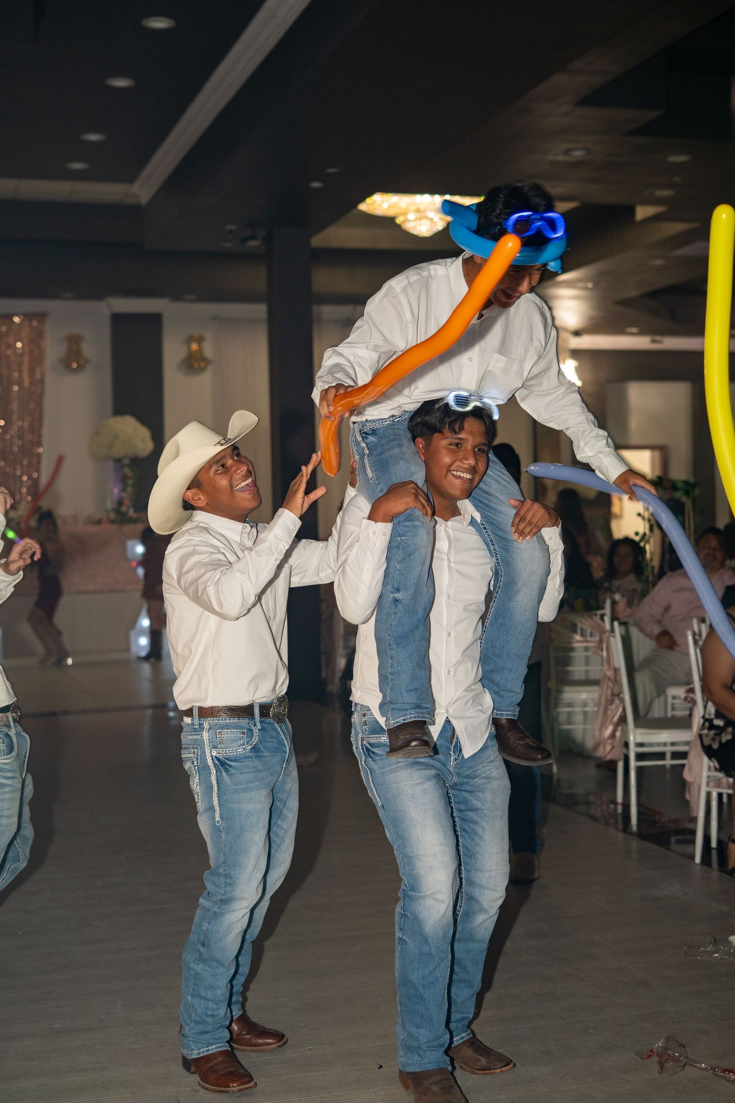 Men in cowboy attire lift a child at a festive quinceañera celebration with colorful balloon decorations.