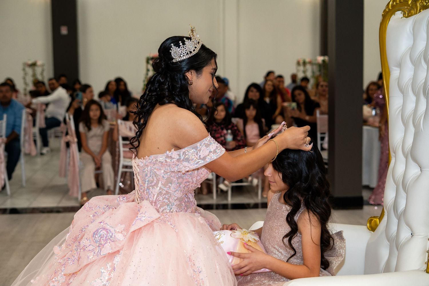 A quinceañera in a pink gown places a tiara on a younger girl at her party.
