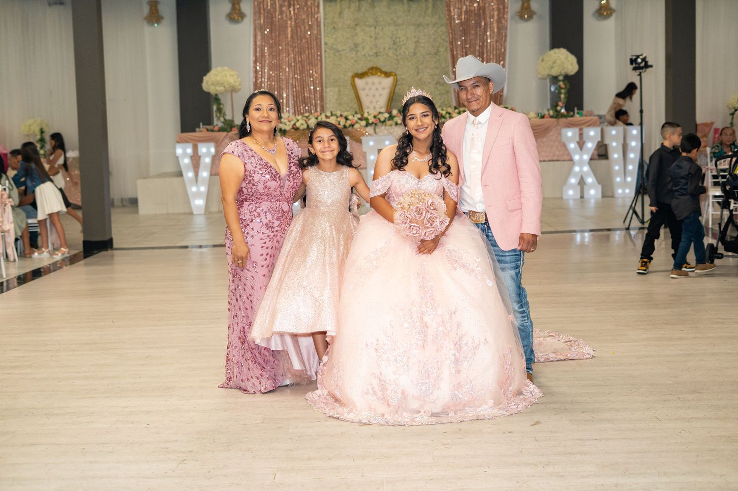 Quinceañera girl in pink gown poses with family including parents and young sister on dance floor.