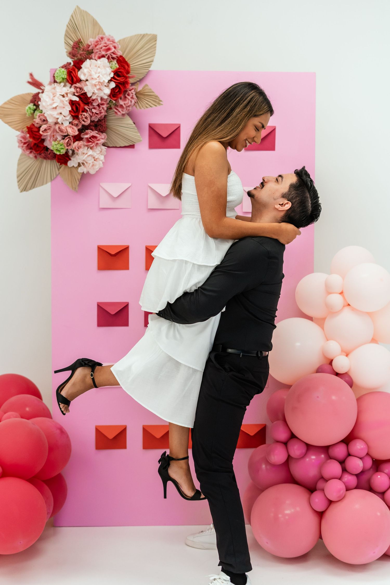 Couple embracing in romantic lift pose before pink wall with colorful envelopes and balloon decorations.