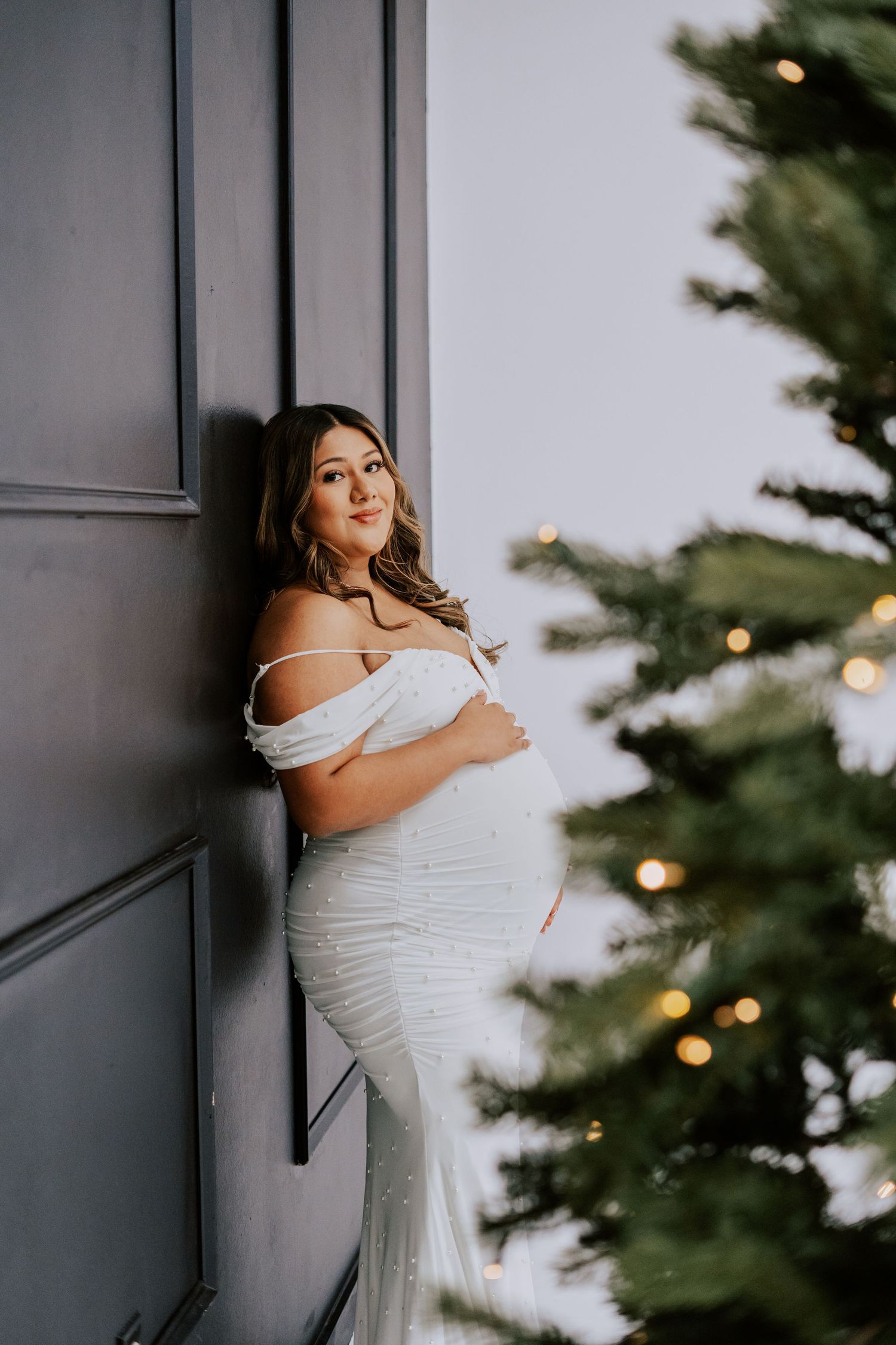 Pregnant woman in white off-shoulder gown leaning against wall near bokeh Christmas tree lights.