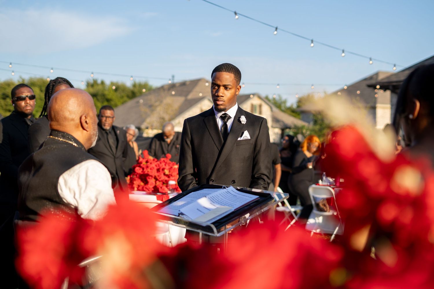 Groom in black suit receives wedding rings at outdoor ceremony surrounded by vibrant red floral arrangements.
