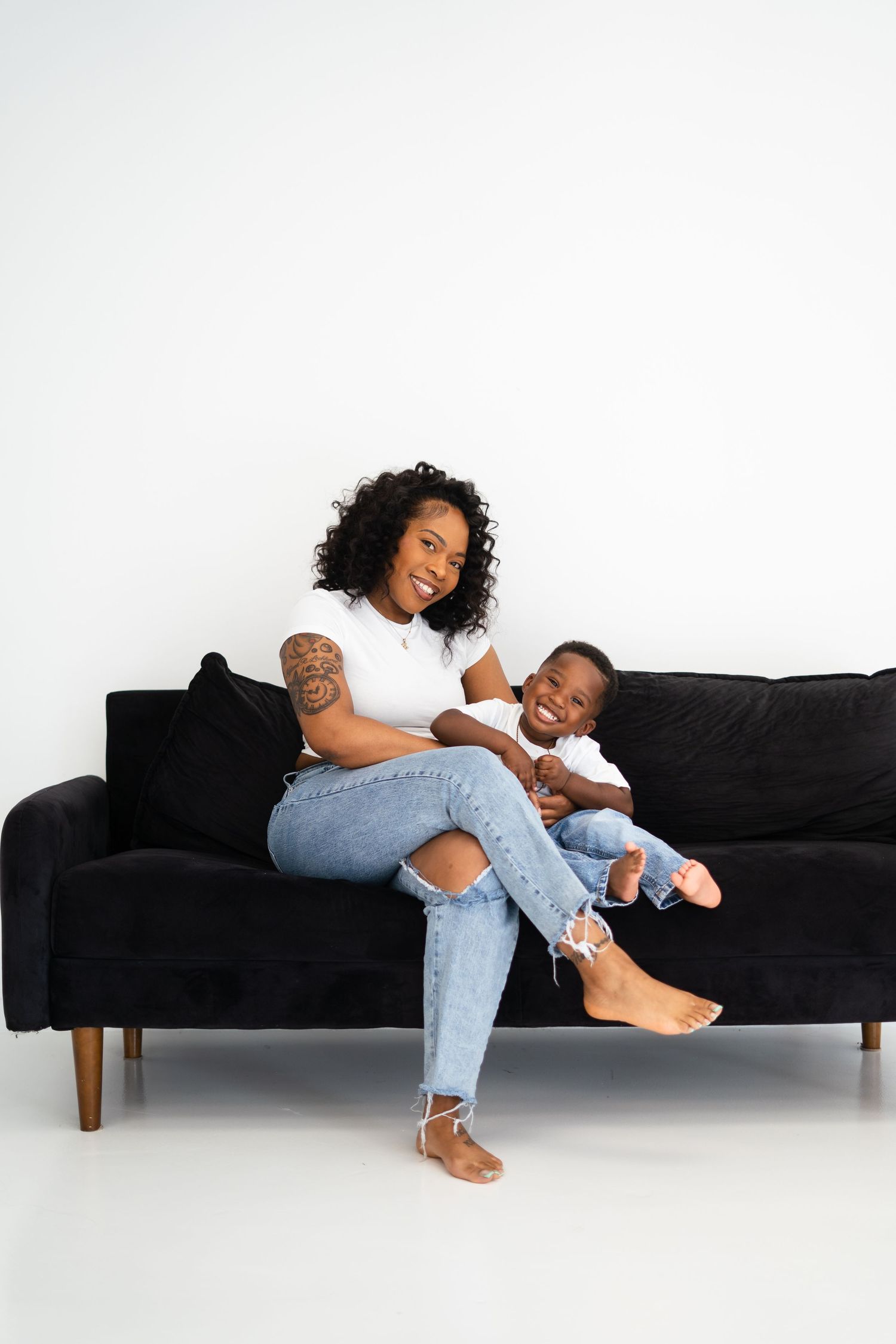 A mother sits on a black sofa holding her smiling toddler, both wearing white tops and jeans.