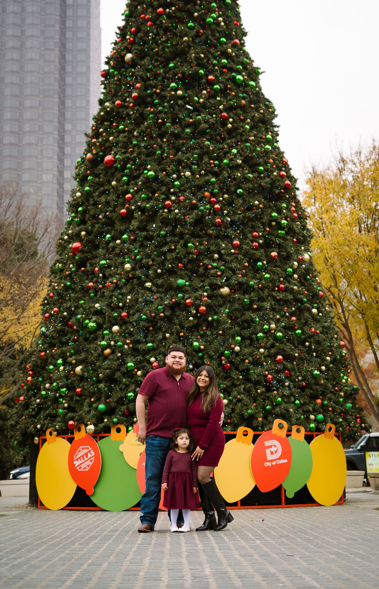 Family of three in matching burgundy outfits posing in front of a giant decorated Christmas tree outdoors.